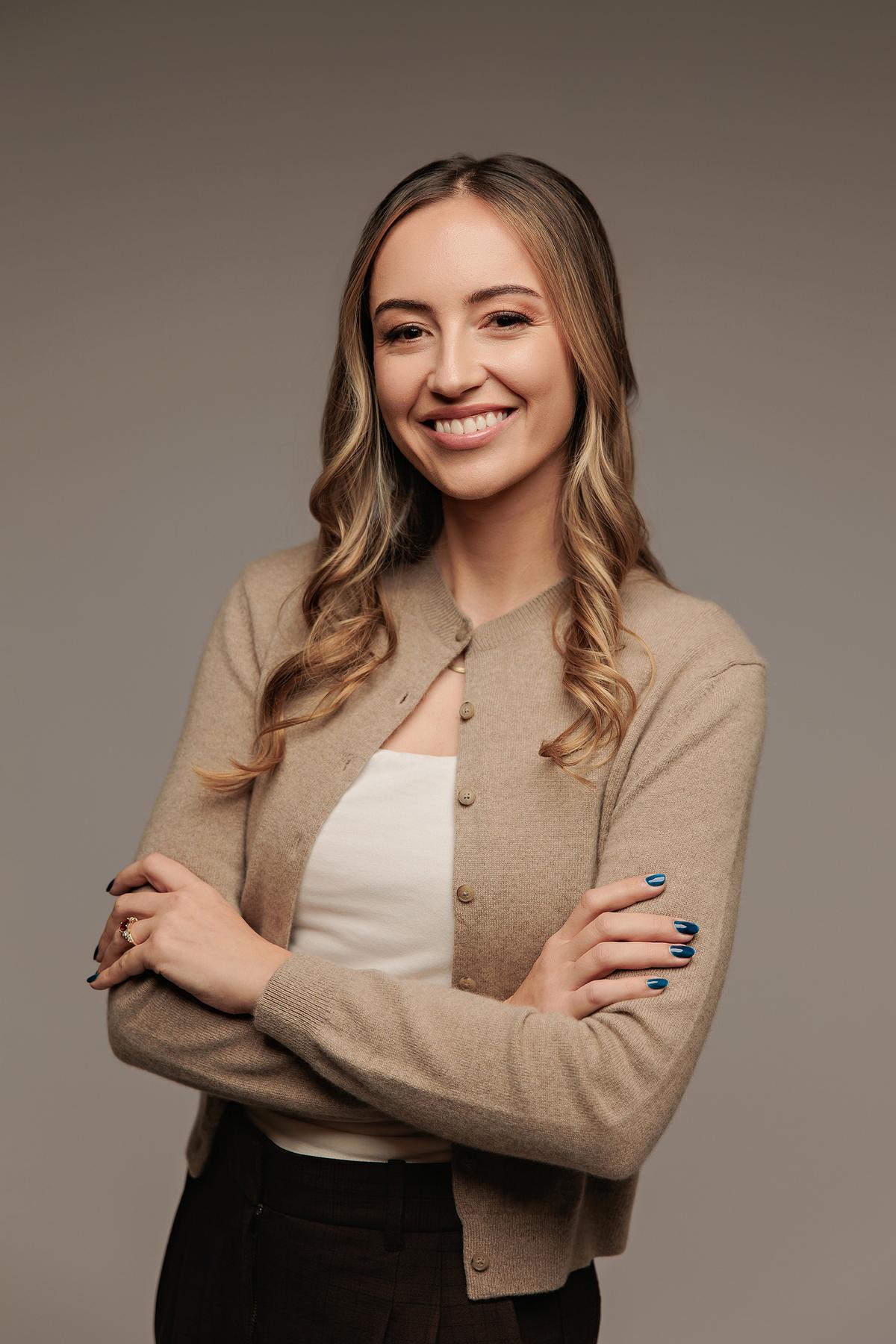 Professional headshot Calgary studio: Bright smiling young woman in a tan cardigan and white top with arms crossed against a soft grey background