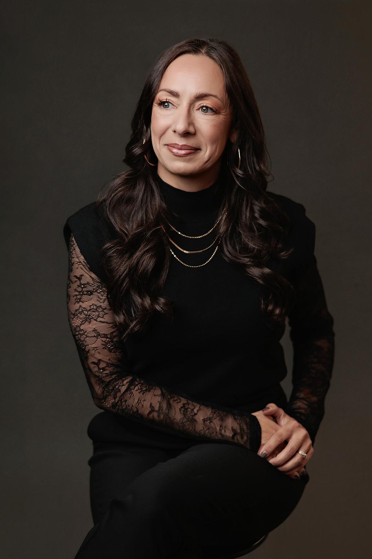 A woman with long dark hair wears a black lace-sleeved top in this professional Calgary headshot, captured with soft studio lighting against a dark backdrop.