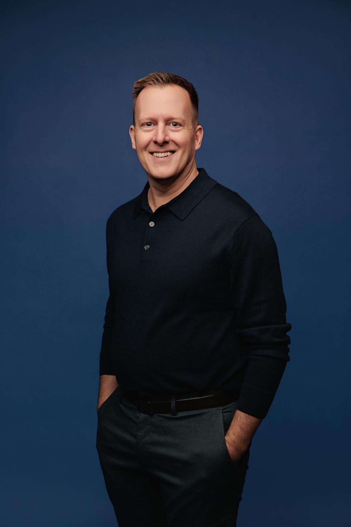 Professional headshot Calgary studio: Smiling man in a black long-sleeve polo with hands in pockets against a rich deep blue studio background