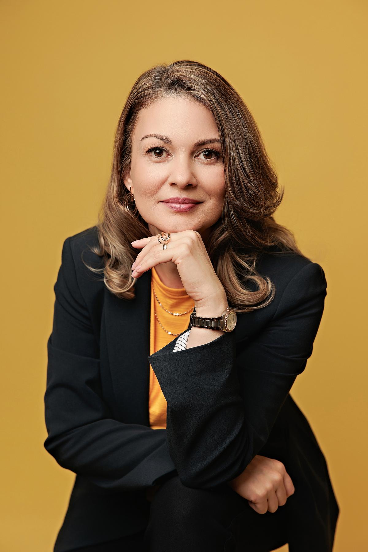Personal branding headshot Calgary studio: Confident businesswoman in a black blazer and orange top with chin resting on hand and a thoughtful expression, warm golden amber background