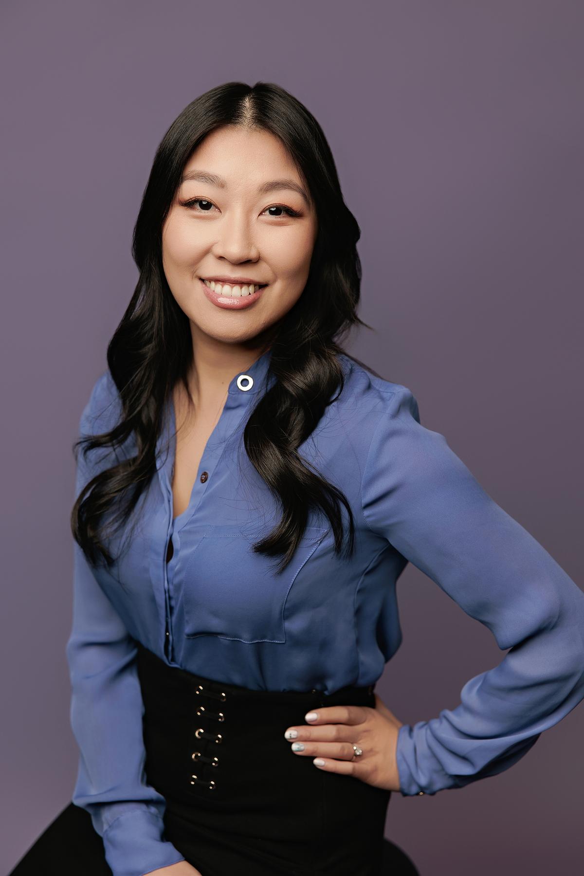 Professional headshot Calgary studio: Confident young woman in a cornflower blue silk blouse and black lace-up corset skirt with hand on hip and a bright smile, soft lavender background