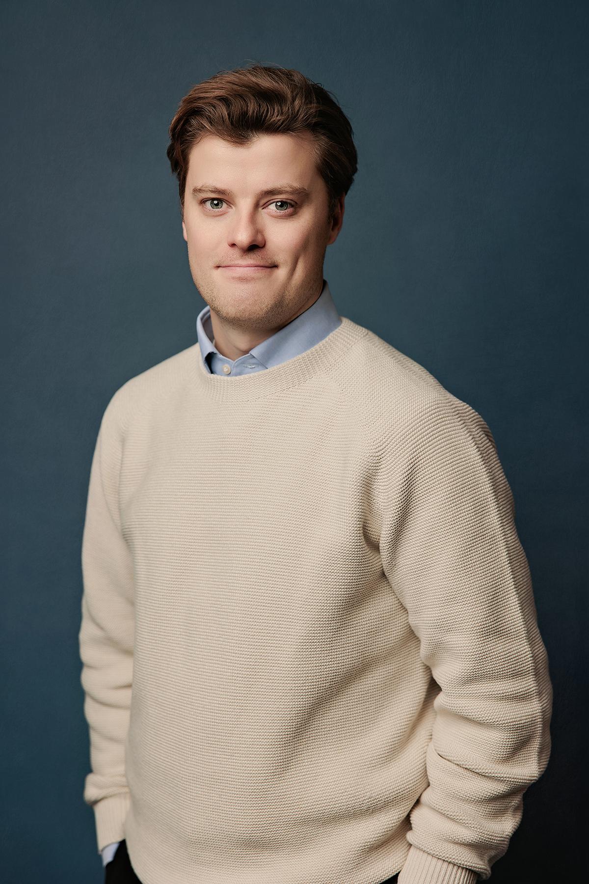 Team headshots Calgary studio: Approachable young male professional with auburn hair in a cream textured knit sweater over a light blue shirt standing with hands in pockets against a deep teal studio background