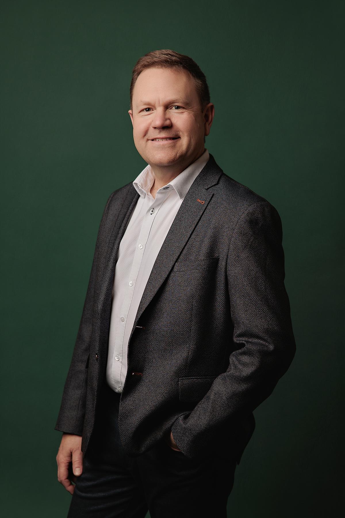 Professional headshots Calgary studio: Confident male executive with short salt-and-pepper hair in a dark grey fleck blazer with orange accent stitching and a white dress shirt, standing with hand in pocket and a warm smile against a rich emerald green background