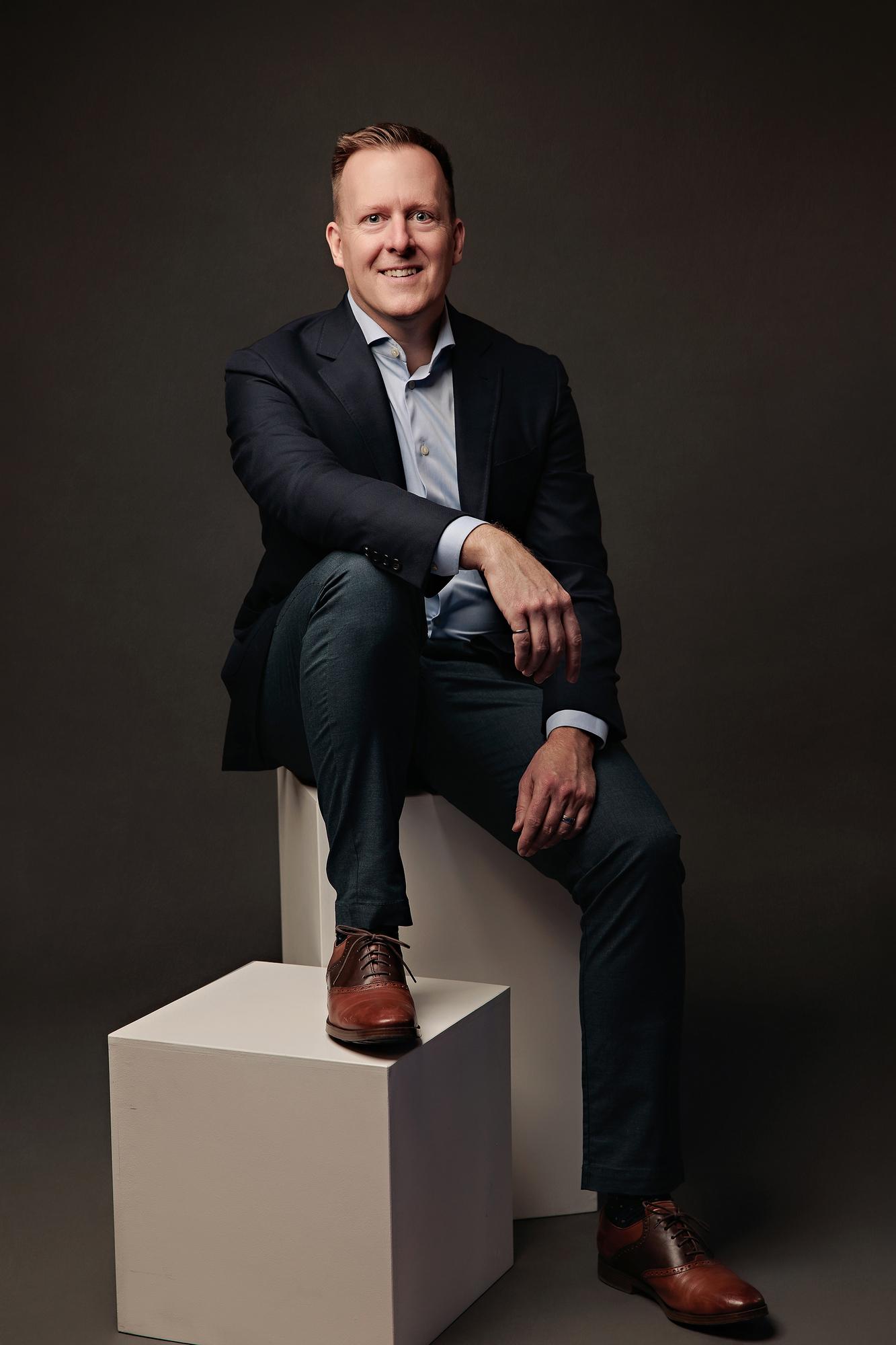 Executive headshot Calgary studio: Professional man in a navy blazer and light blue shirt seated on a white cube with one foot elevated, dark background