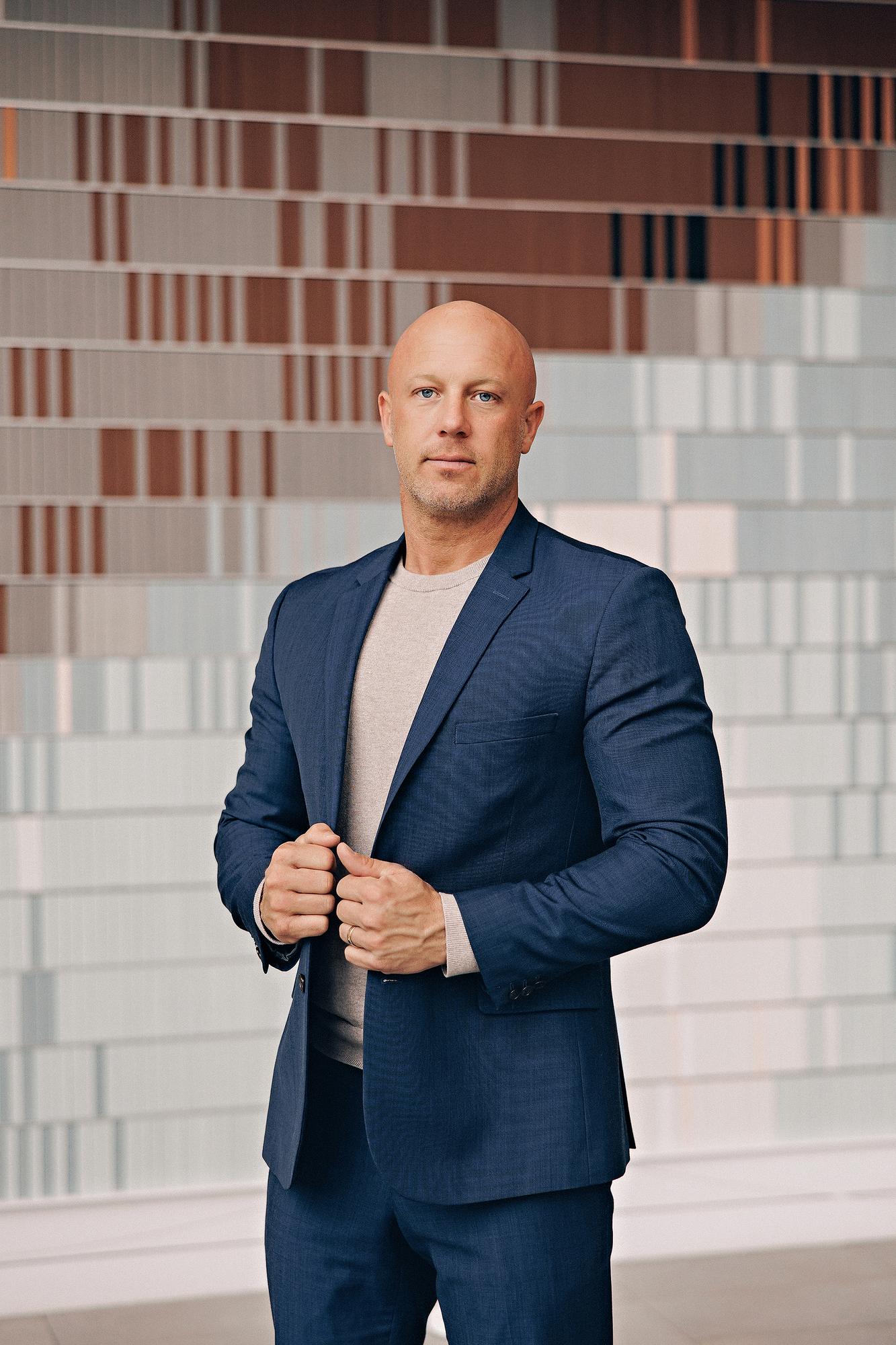 Personal branding photography Calgary: Powerful professional man in a navy blue blazer and grey t-shirt adjusting his jacket with both hands in front of a colourful striped feature wall inside a modern Calgary building