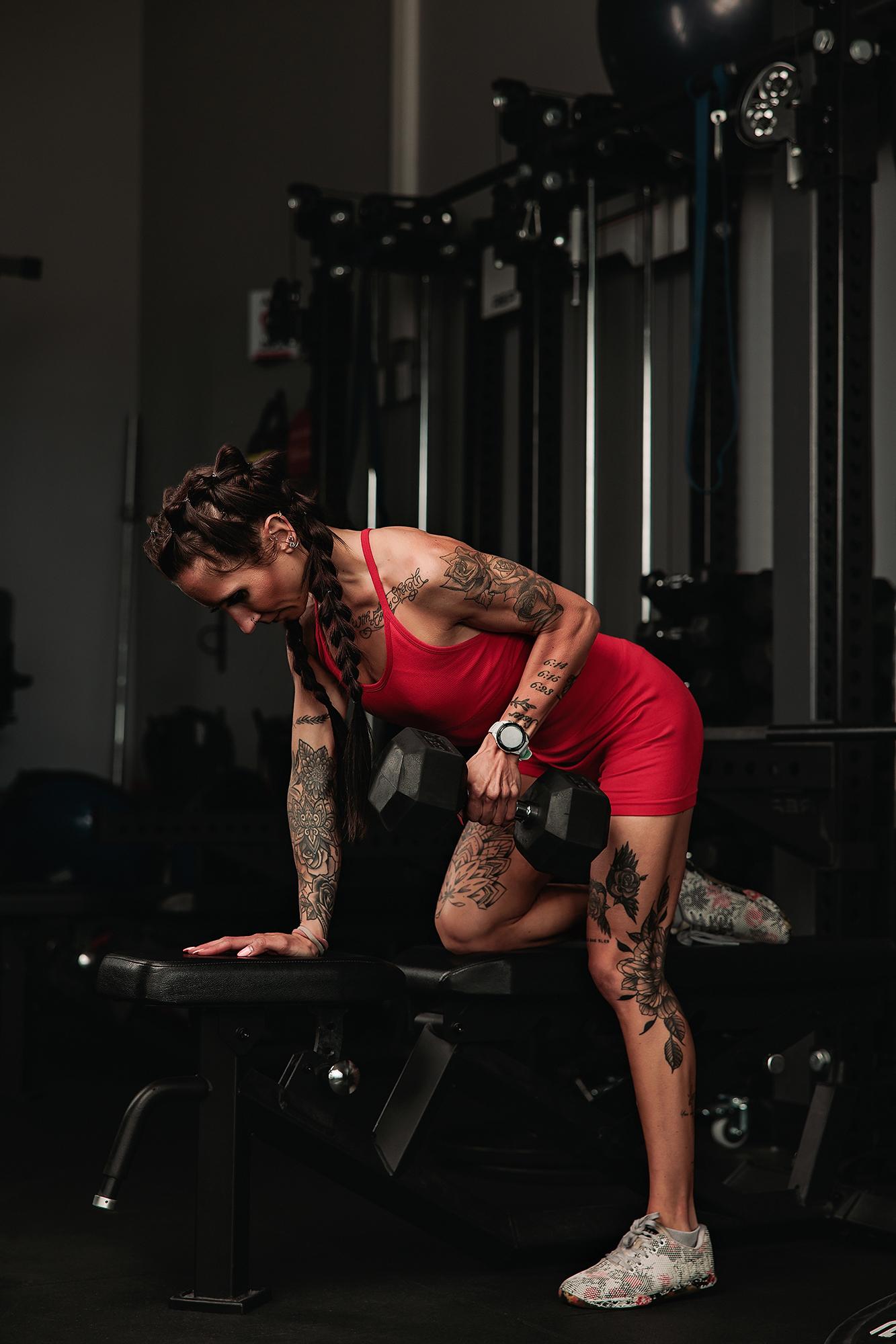Personal branding photography Calgary gym: Tattooed female athlete in a red sport set performing a single-arm dumbbell row on a bench, heavy gym equipment and cable machines behind her