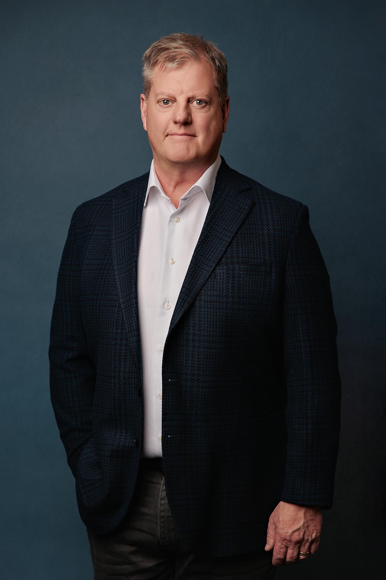Team headshots Calgary studio: Authoritative senior male executive in a dark navy herringbone blazer and white dress shirt standing with hands in pockets against a deep teal studio background