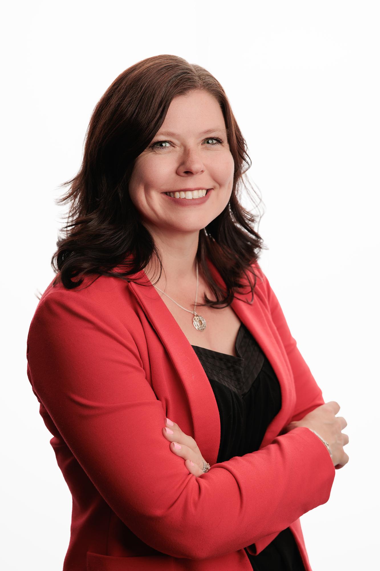 Team headshots Calgary studio: Confident professional woman with dark auburn wavy hair in a red blazer over a black top with arms crossed and a silver pendant necklace, white background