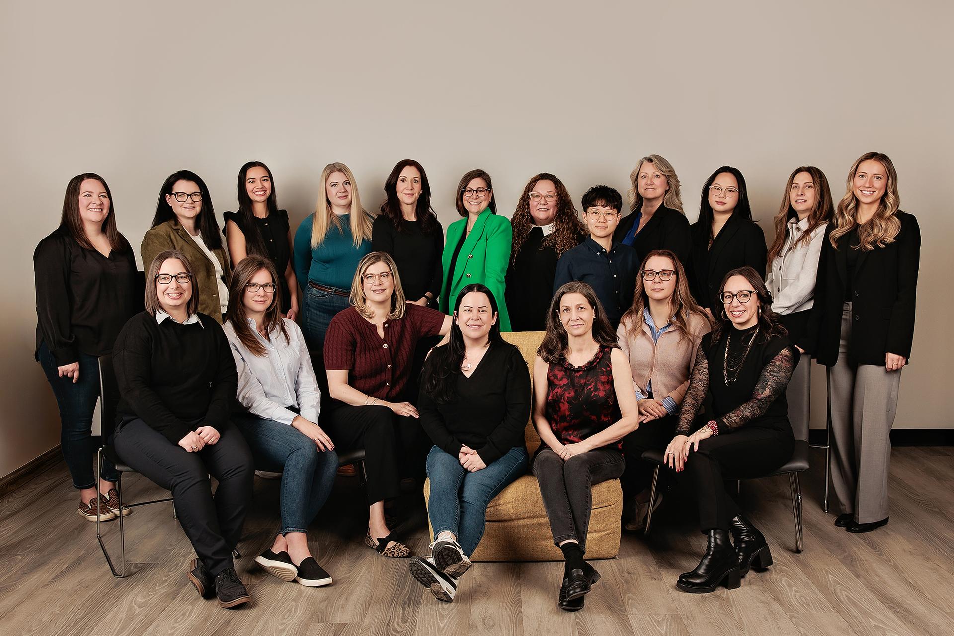 Corporate group team photography Calgary studio: Large team of nineteen women in two rows — standing and seated — against a warm taupe studio backdrop, showcasing diverse professional attire including a standout green blazer