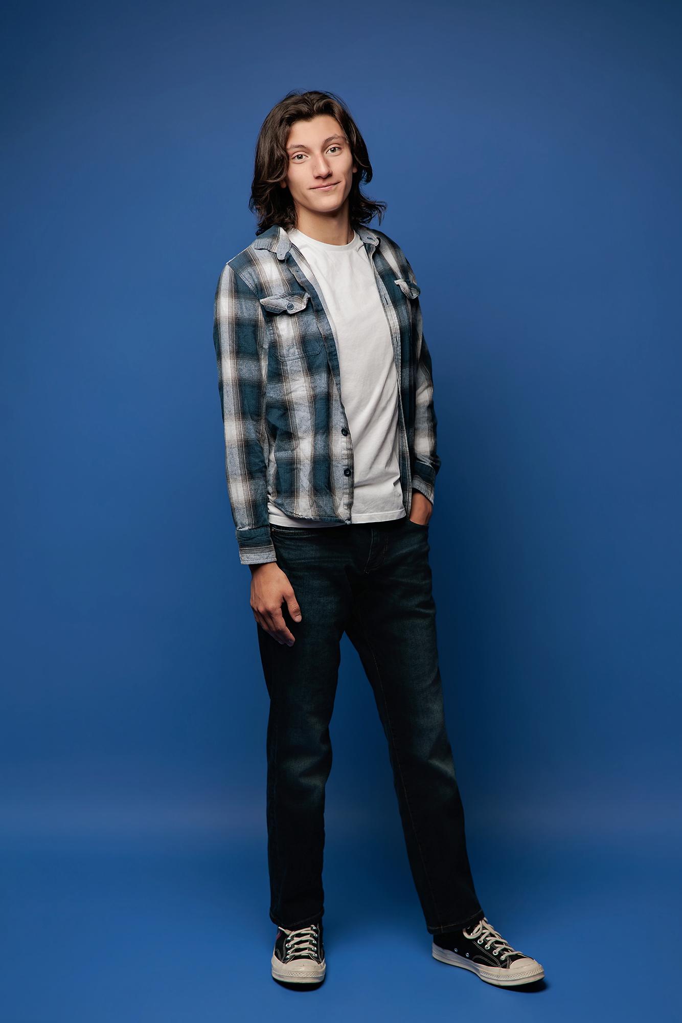 A young man with long hair wears a plaid shirt and jeans in this Calgary studio headshot, showcasing a relaxed pose against a vibrant blue backdrop.