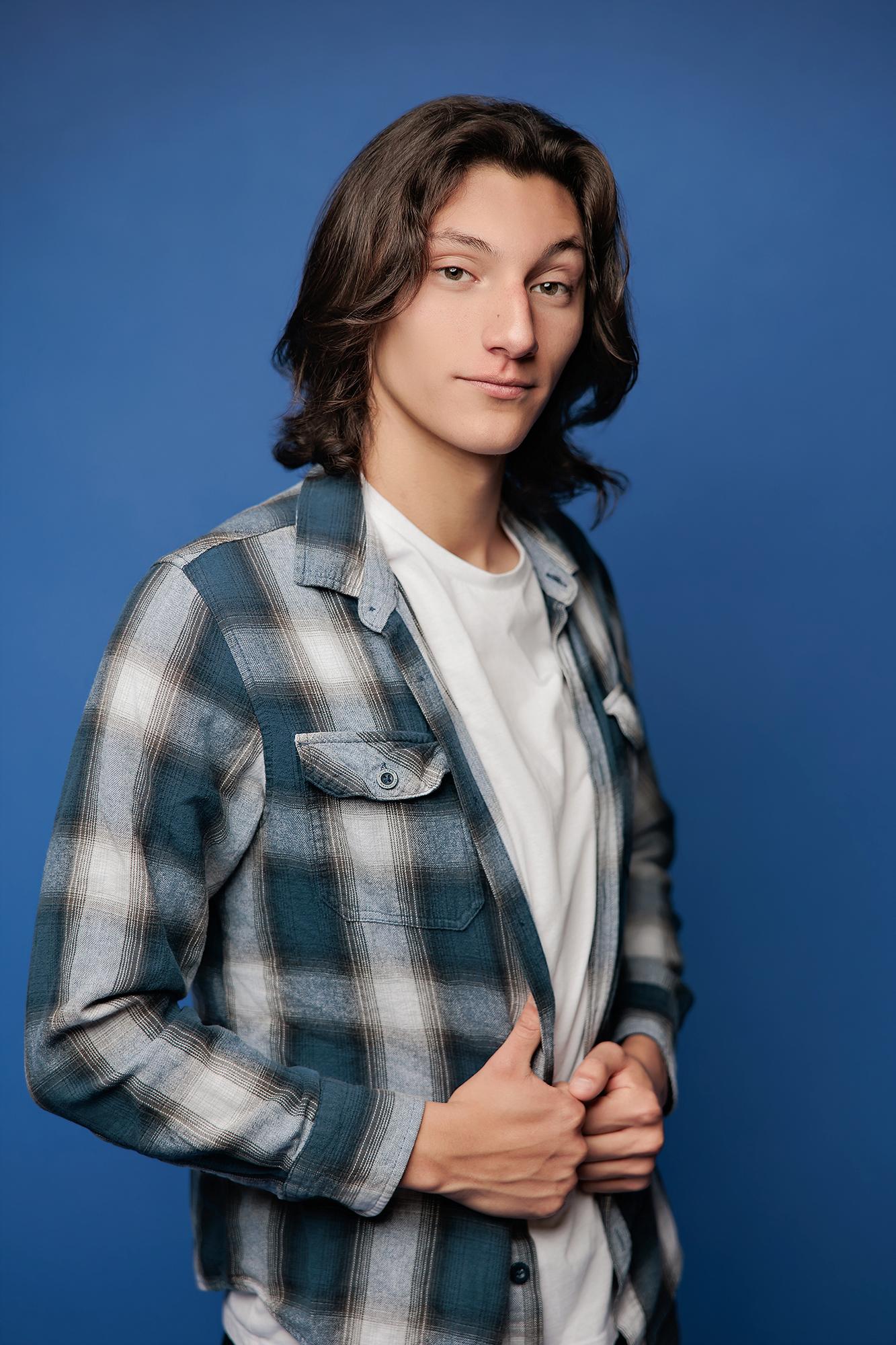 A young man with long brown hair wears a plaid shirt in this Calgary headshot session, captured with soft studio lighting against a blue backdrop.