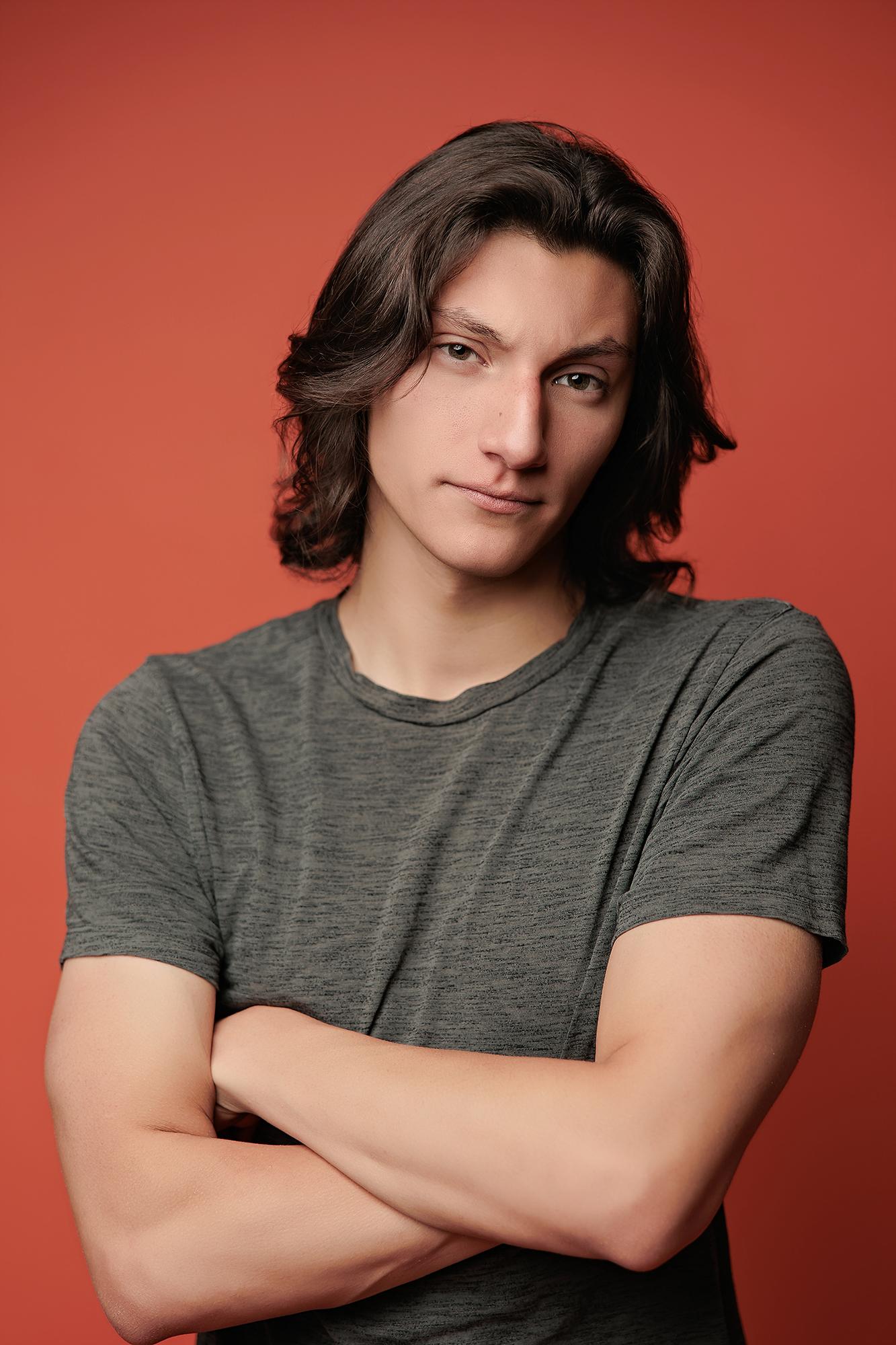 A young man with long dark hair poses with crossed arms in this Calgary photography studio portrait, featuring soft lighting against a bold red backdrop.