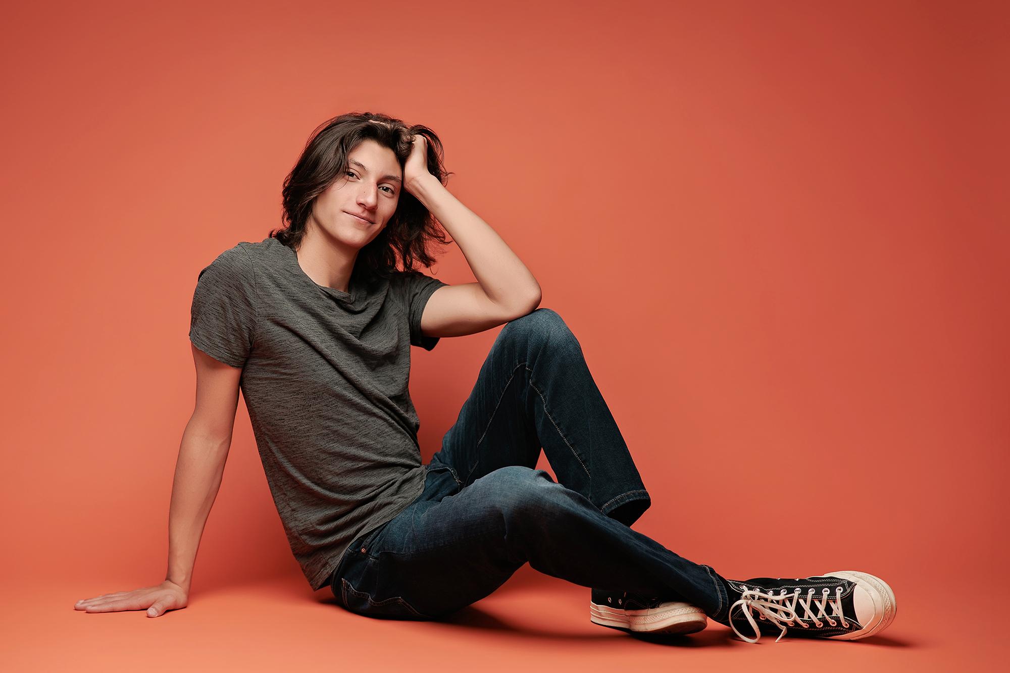 A young man with long hair sits casually in a Calgary photography studio, wearing a grey t-shirt and jeans against a vibrant coral backdrop.