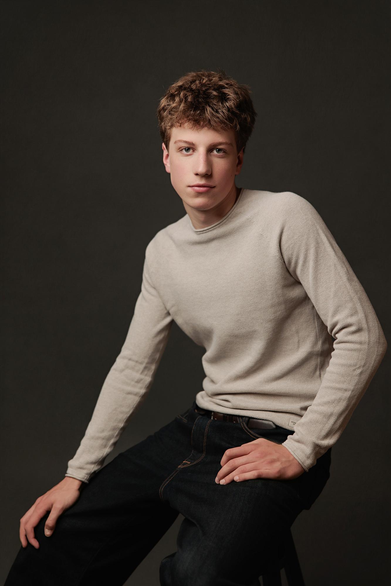 Young actor with curly hair wearing a beige sweater sits for a professional actor portrait at our Calgary photography studio, featuring soft studio lighting.