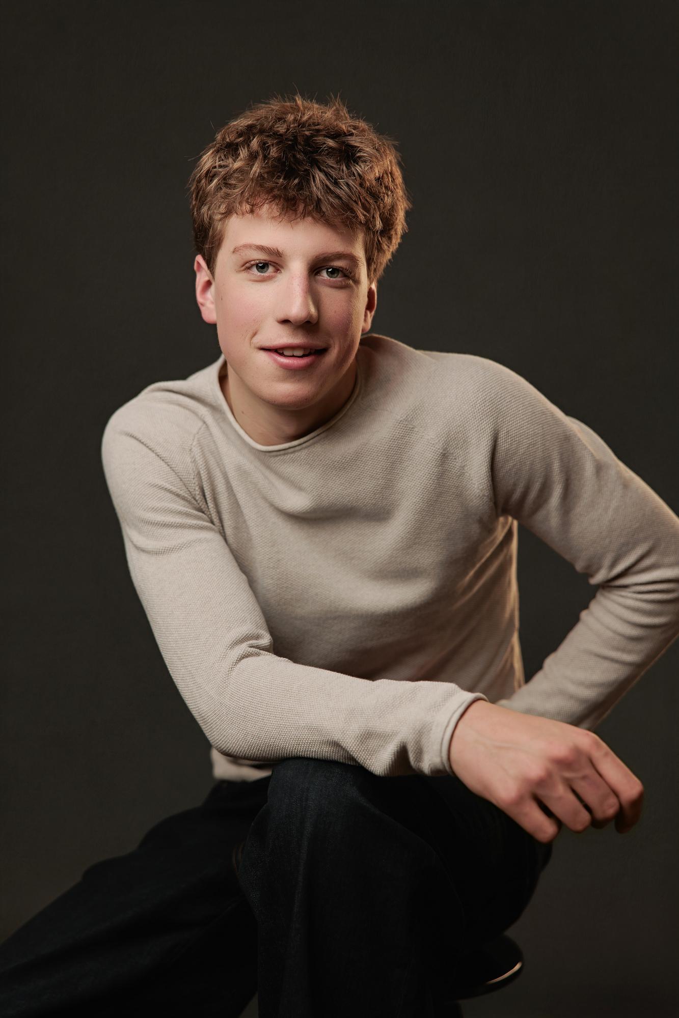 A young actor with textured brown hair wears a beige sweater in this Calgary actor headshot session, featuring soft studio lighting against a dark backdrop.