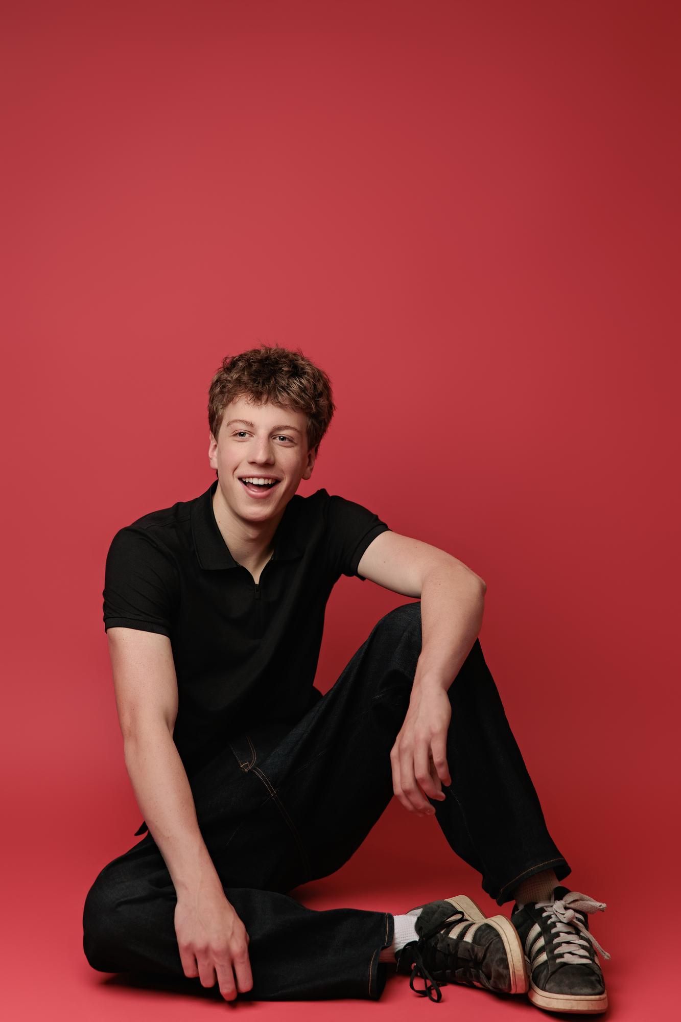 A young actor with curly hair wearing a black polo sits against a vibrant red backdrop, showcasing the modern style of a Calgary actor headshot photographer.