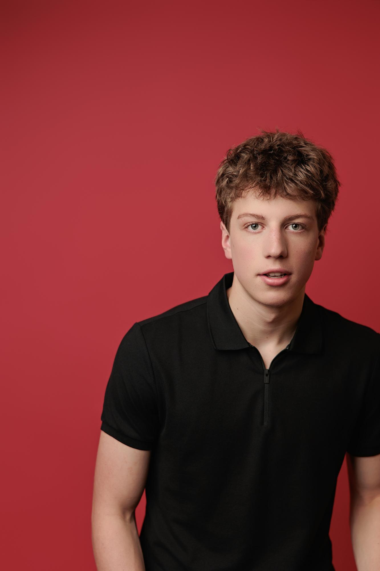Young actor with messy hair wears a black polo shirt against a vibrant red backdrop, captured by our Calgary actor headshot photographer for a modern look.
