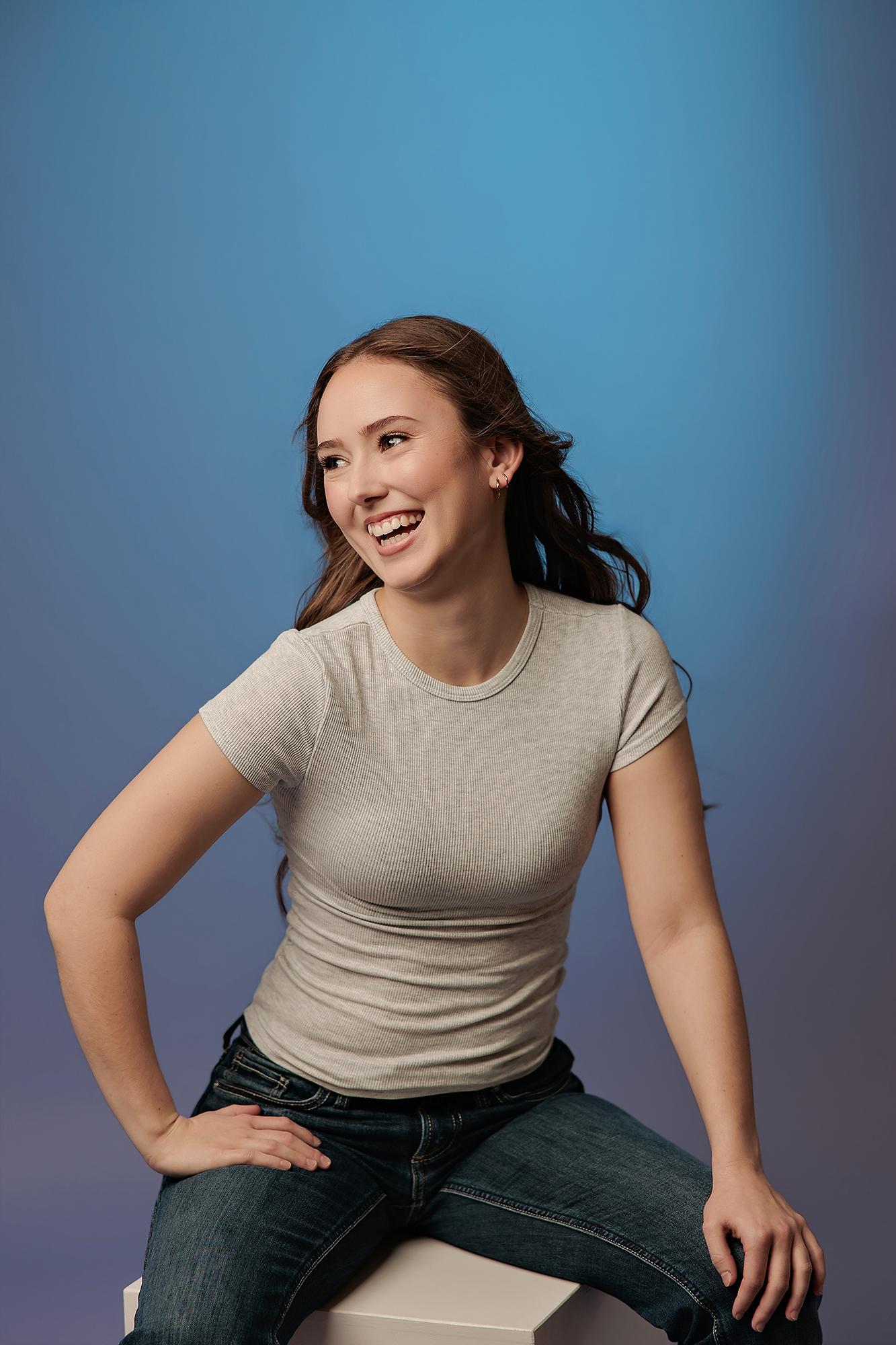 A smiling woman in a casual grey t-shirt poses for a Calgary actor headshot photographer, showcasing a relaxed and authentic expression against a blue studio backdrop.
