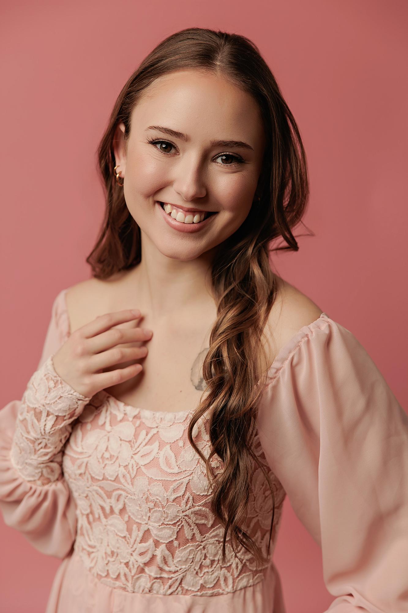 A smiling young woman in a lace-detailed pink dress poses for a Calgary actor headshot photographer, showcasing soft lighting and a warm studio aesthetic.
