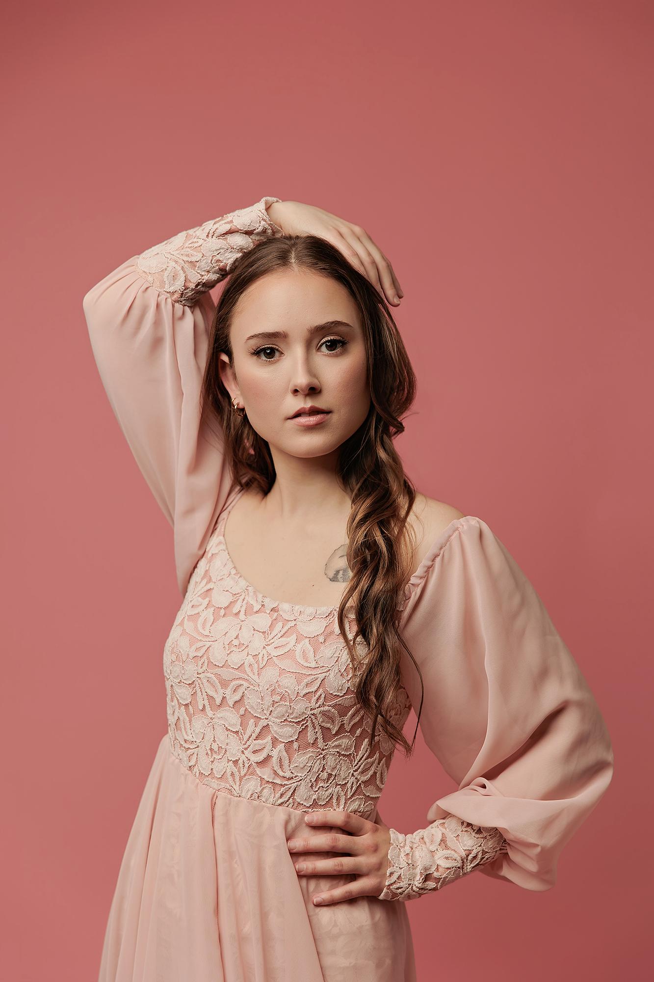 A woman in a lace-detailed blush dress poses gracefully for this Calgary actor photography studio session, showcasing soft lighting against a solid pink backdrop.