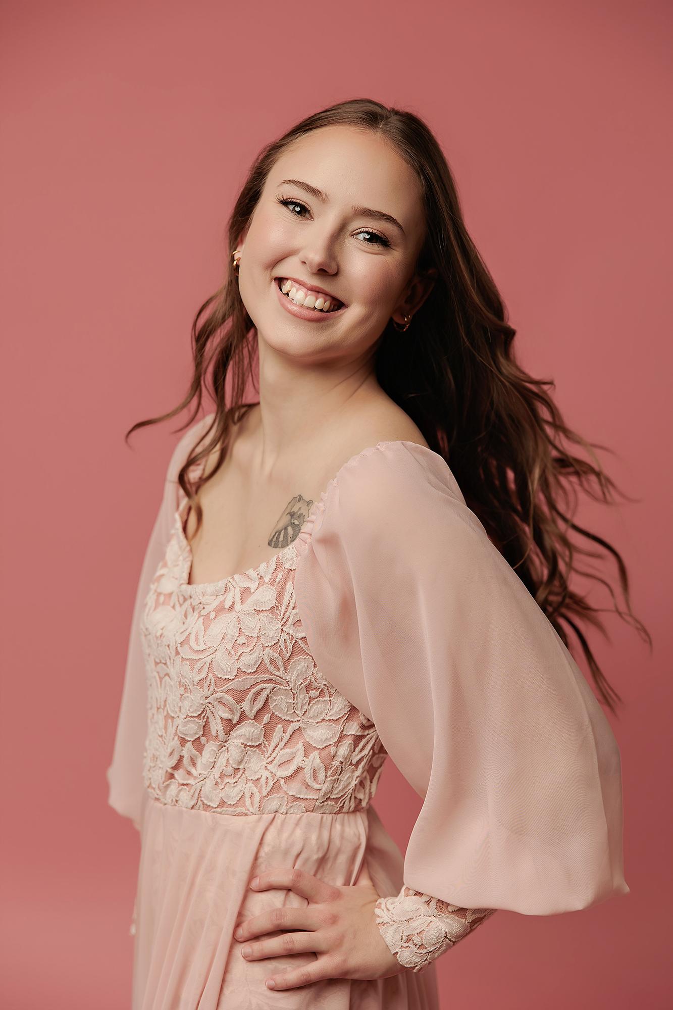 A smiling young woman with long wavy hair wears a pink lace dress in this Calgary photography studio portrait, captured with soft, flattering lighting.