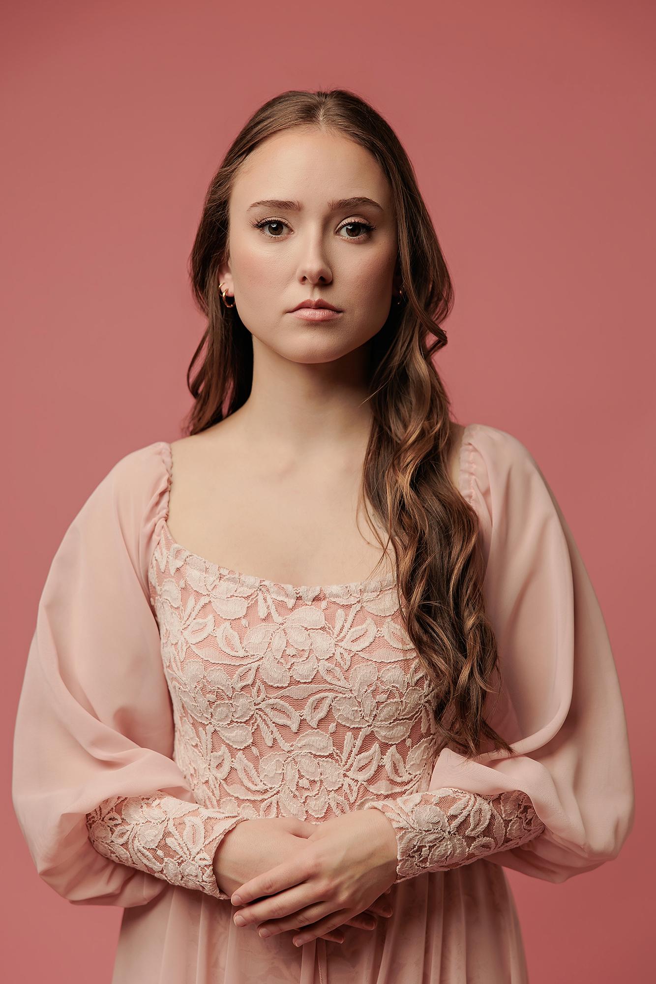 A woman with long wavy hair wears a pink lace dress in this Calgary photography studio portrait, featuring soft lighting and a professional mood.