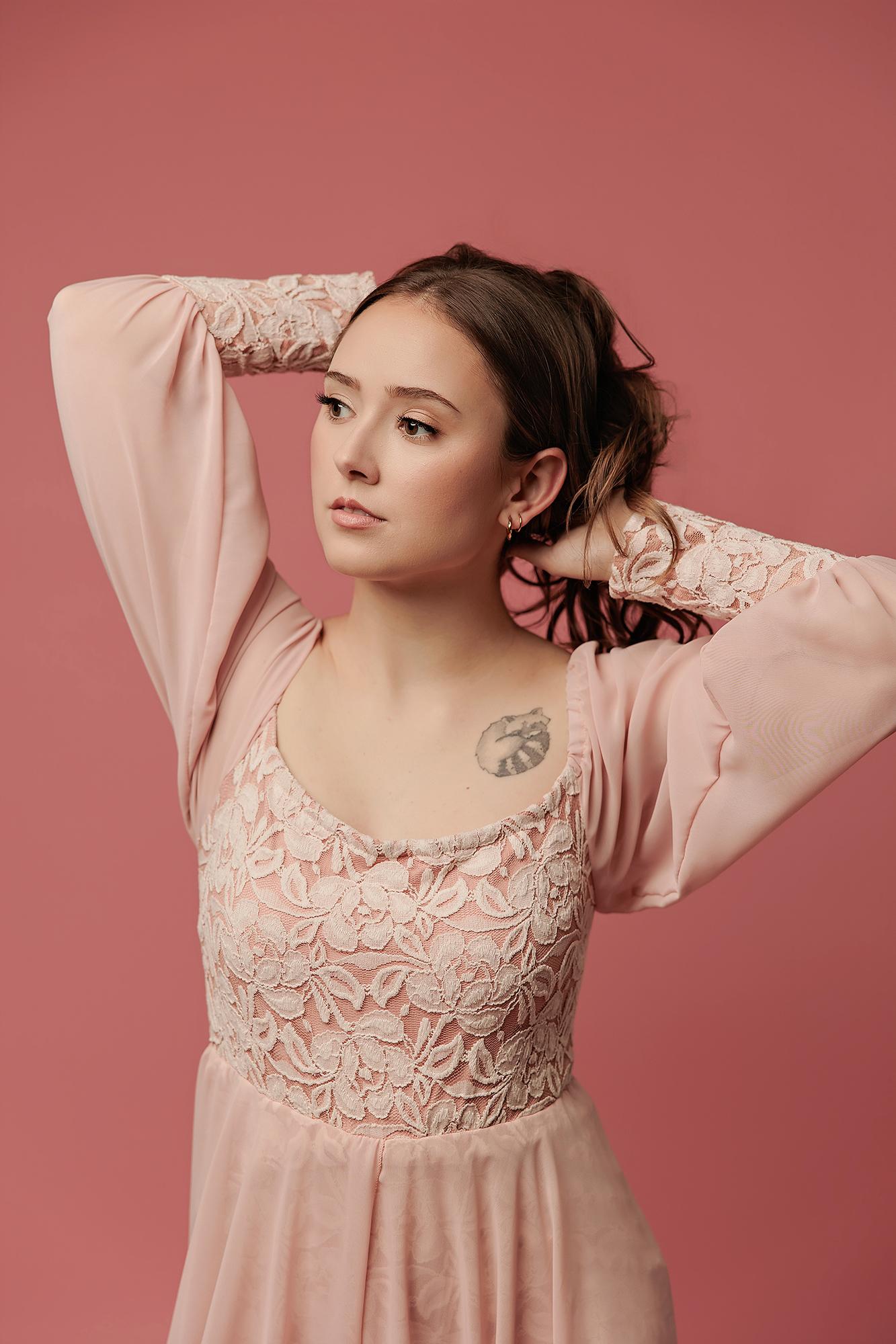 A woman in a lace-detailed blush dress poses for a Calgary photography studio session, her soft expression captured against a matching pink backdrop.