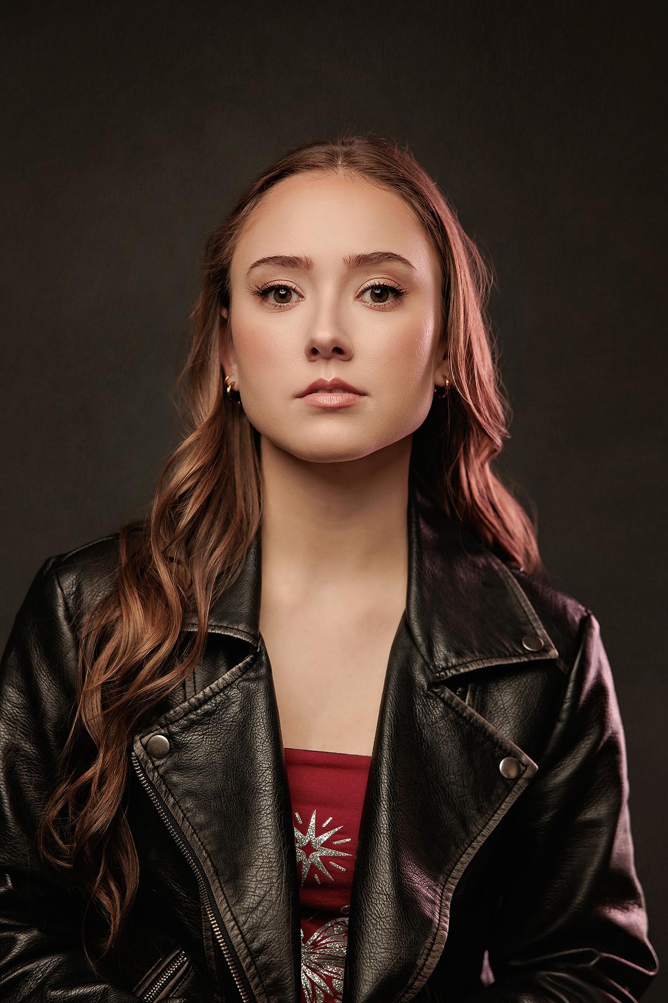 Young woman in a black leather jacket poses for a professional headshot at our Calgary photography studio, featuring moody lighting and a dark backdrop.