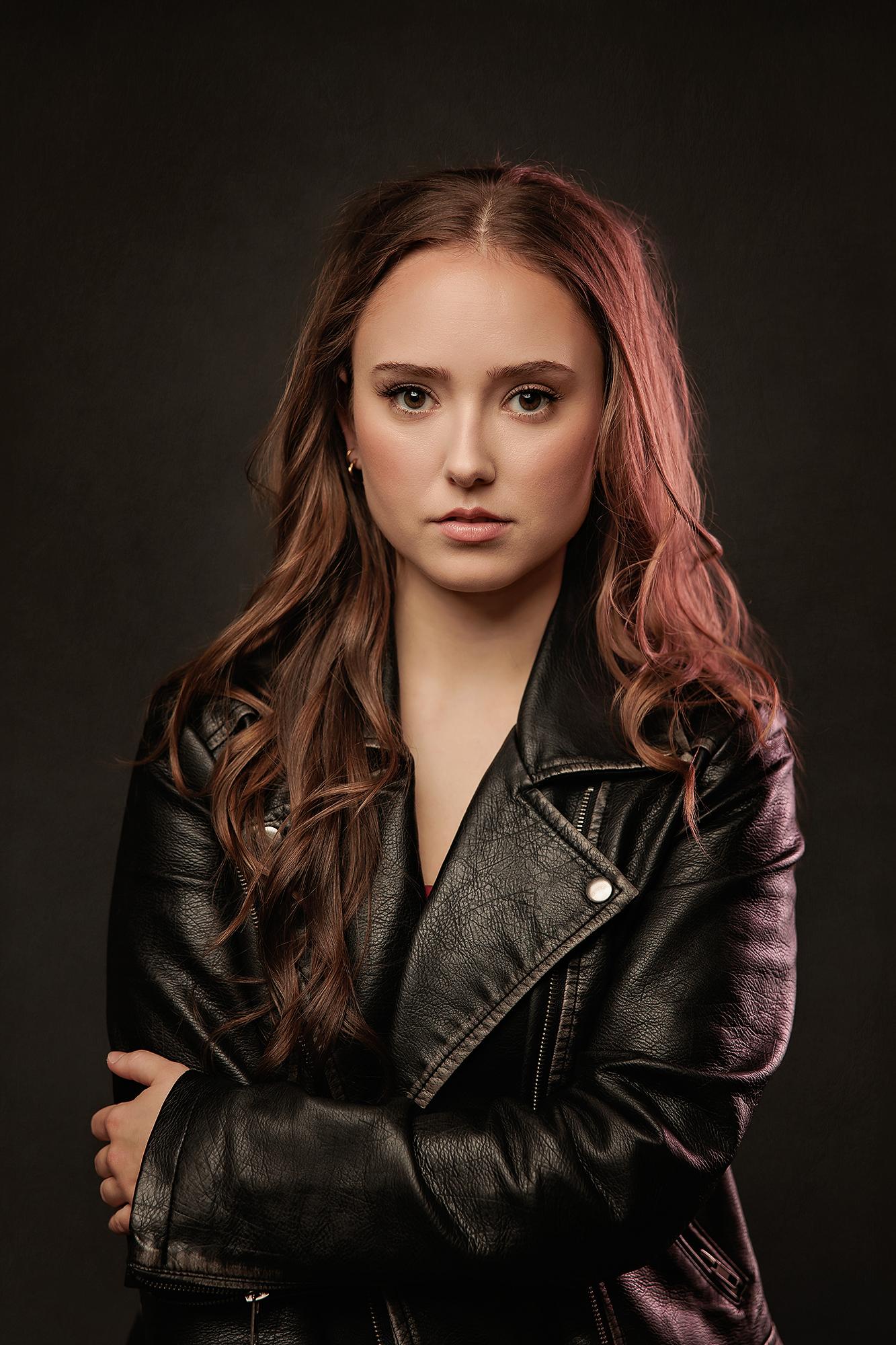 A woman with long wavy hair wears a black leather jacket in this moody Calgary studio headshot, captured with dramatic rim lighting and a serious gaze.