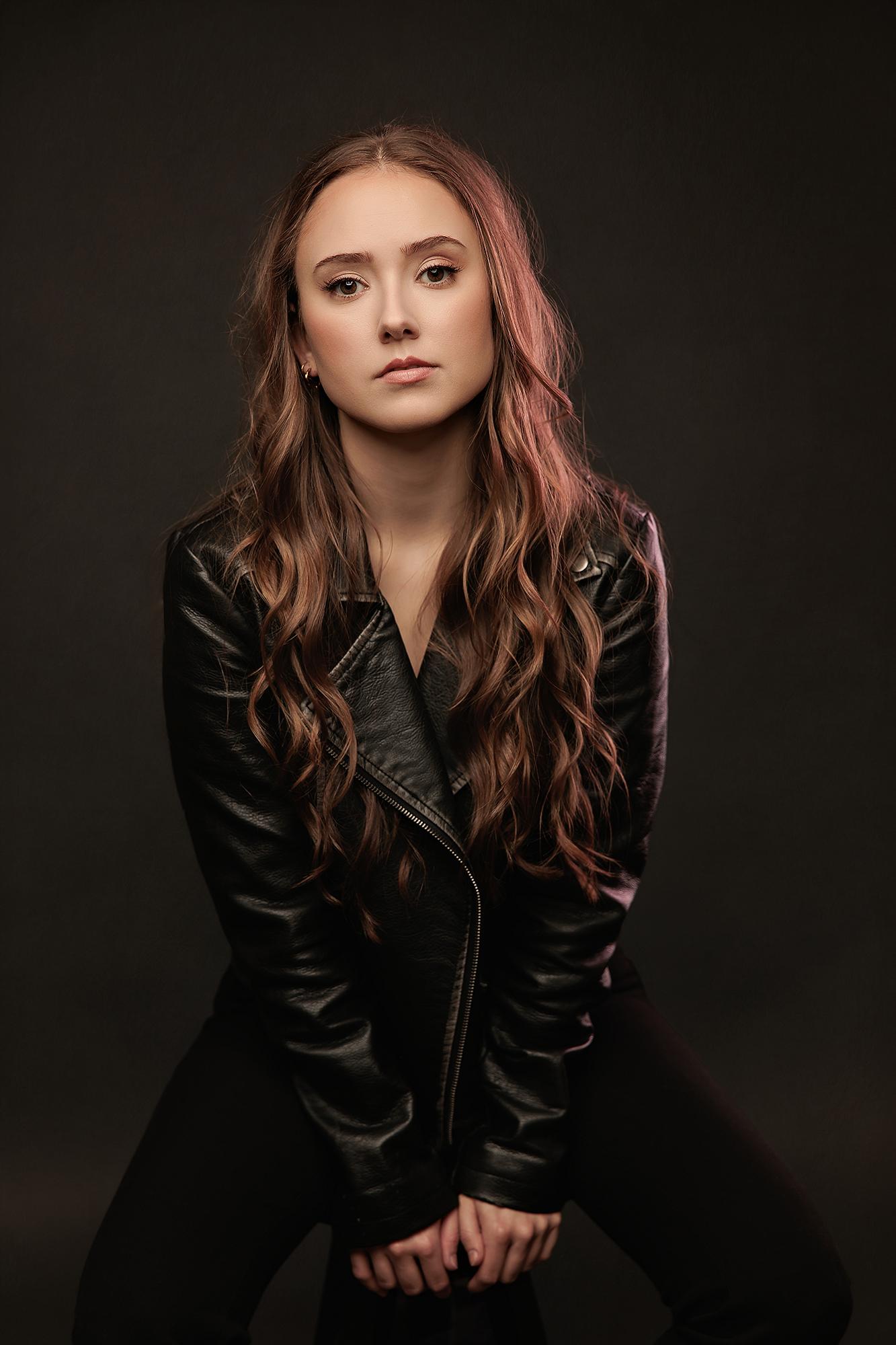 A young woman with long wavy hair wears a black leather jacket in this moody Calgary studio headshot, featuring dramatic lighting and a focused gaze.