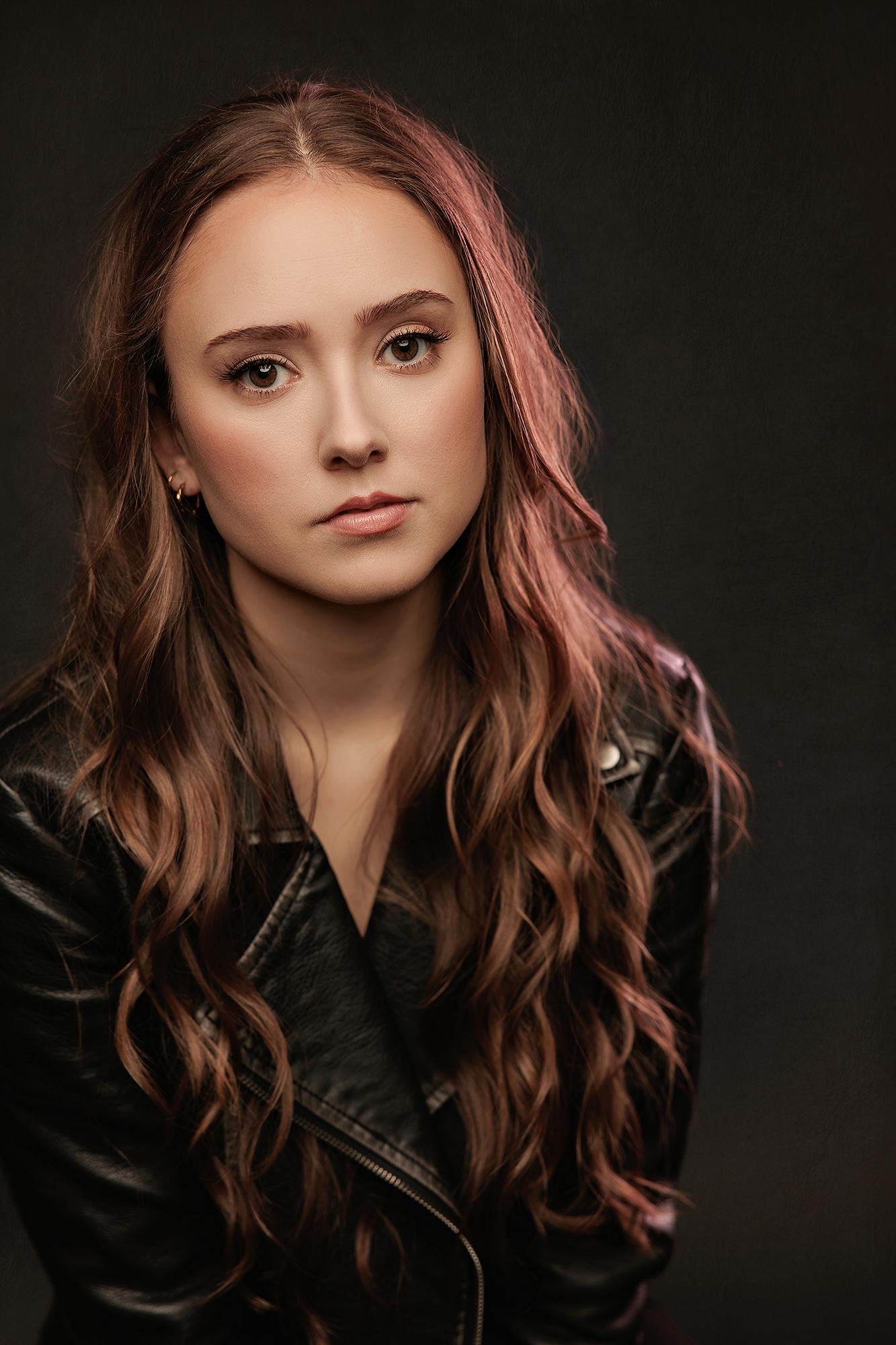 A young woman with wavy hair wears a black leather jacket in this Calgary studio headshot, featuring dramatic rim lighting against a dark backdrop.