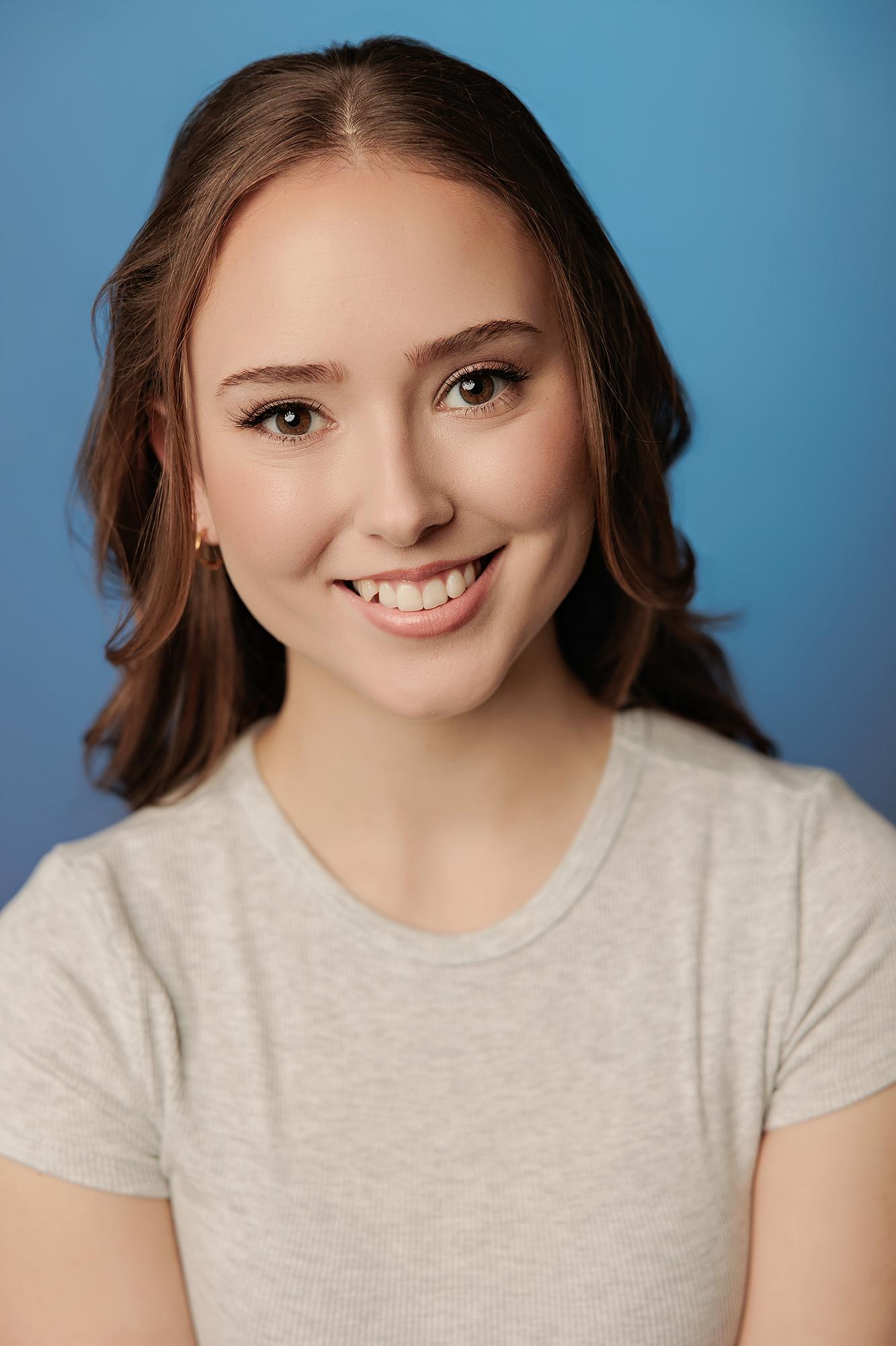 A young actor with a warm smile wears a casual grey shirt against a bright blue background, captured by a professional Calgary actor headshot photographer.