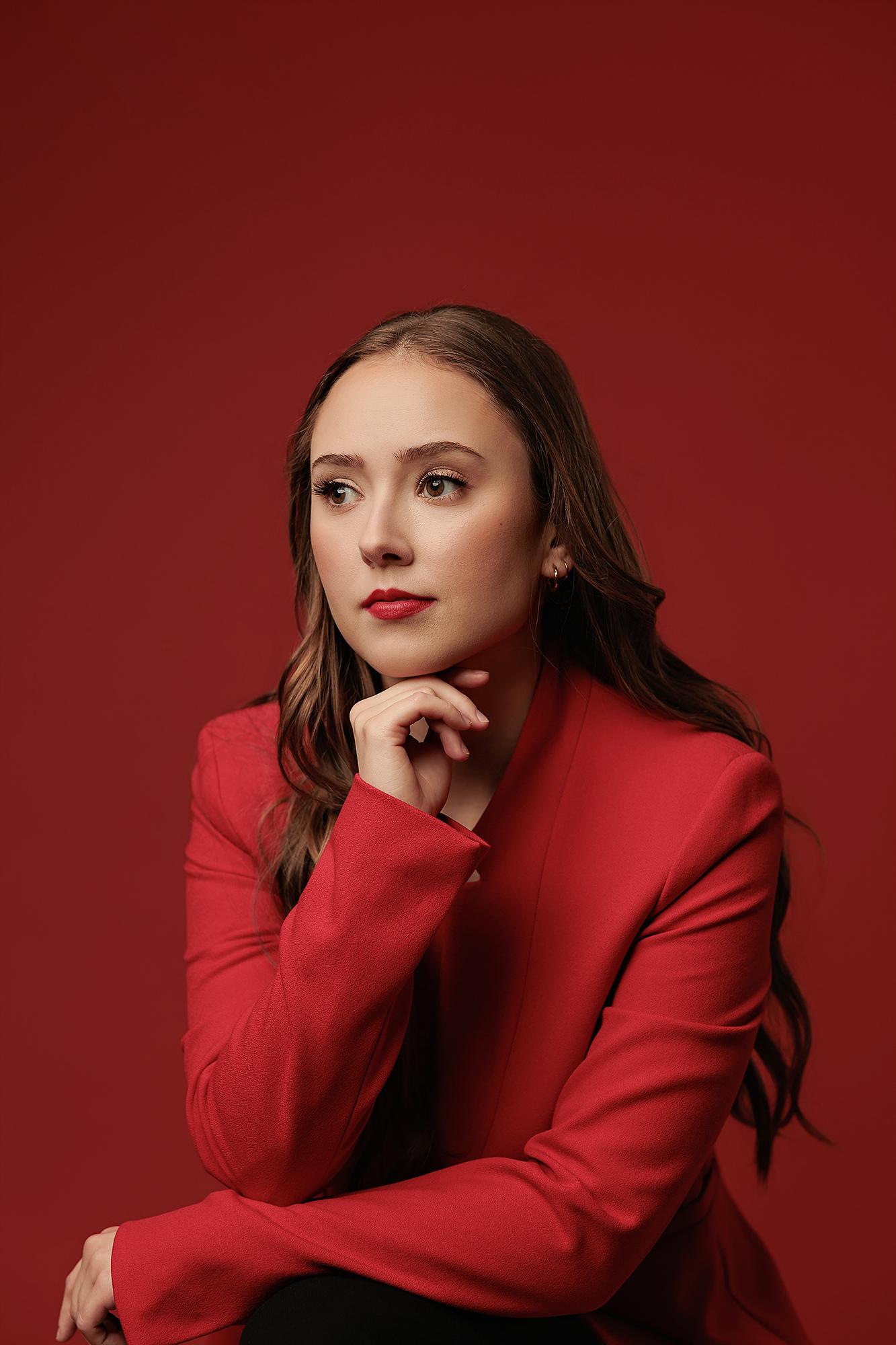 A woman in a red blazer poses thoughtfully for a professional headshot at a Calgary photography studio, featuring soft studio lighting on a red backdrop.