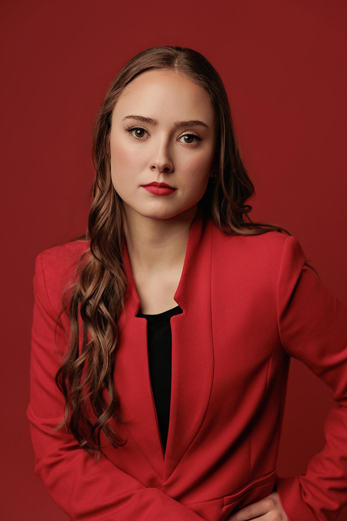 A woman with long wavy hair wears a red blazer in this professional Calgary headshot, captured with studio lighting against a bold matching backdrop.