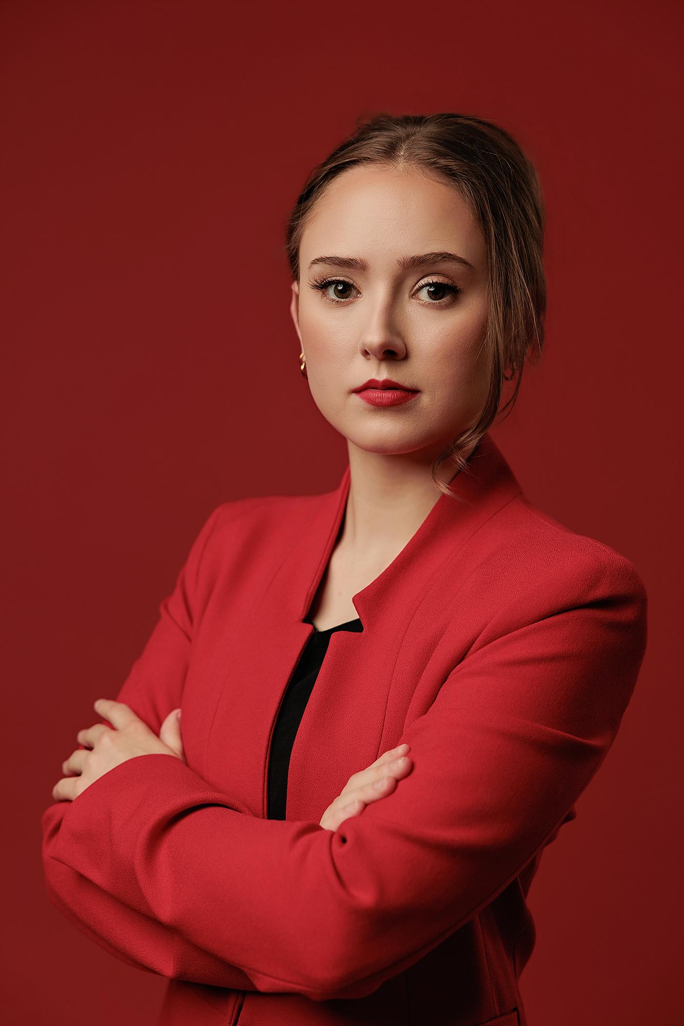 A woman in a red blazer with arms crossed poses for this Calgary headshot session, showcasing professional studio lighting against a bold red backdrop.