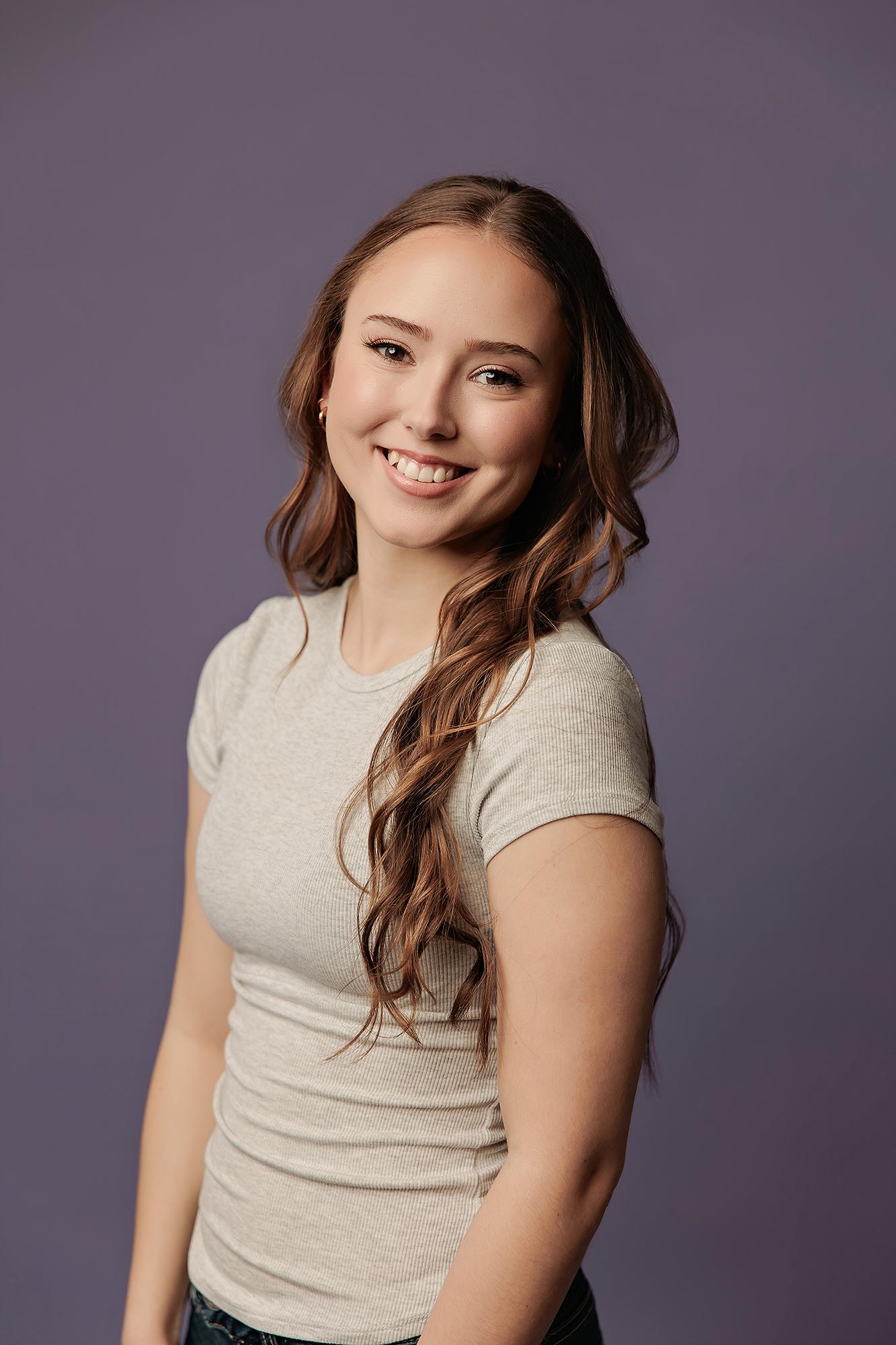 A smiling young woman with long wavy hair wears a casual grey t-shirt in this professional actor headshot captured at a Calgary photography studio.