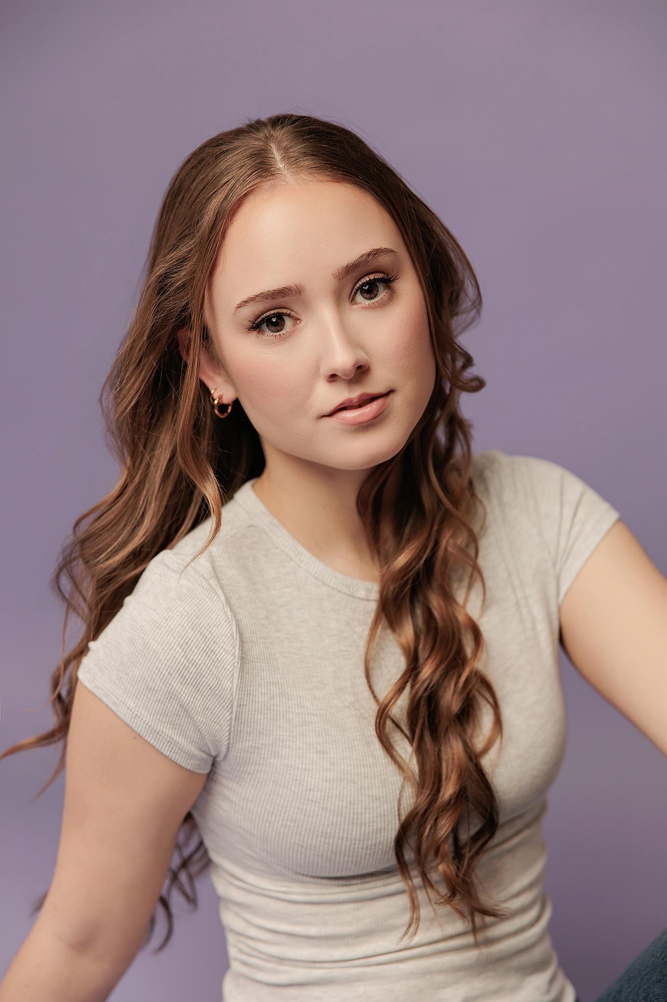 A woman with long wavy hair poses in a grey t-shirt against a purple backdrop, captured by a Calgary actor headshot photographer for a professional look.