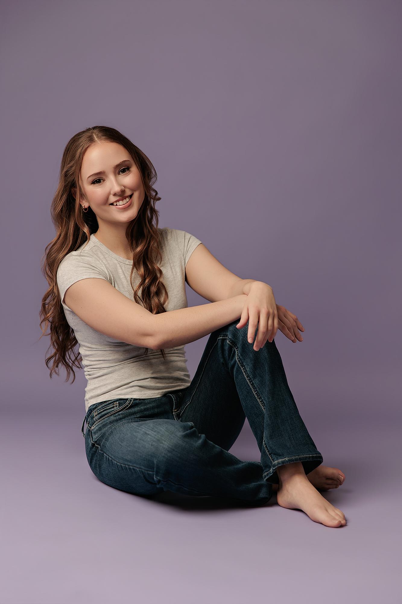 A smiling young woman with long wavy hair sits on the floor in a Calgary photography studio, wearing a casual grey t-shirt and jeans against a purple backdrop.