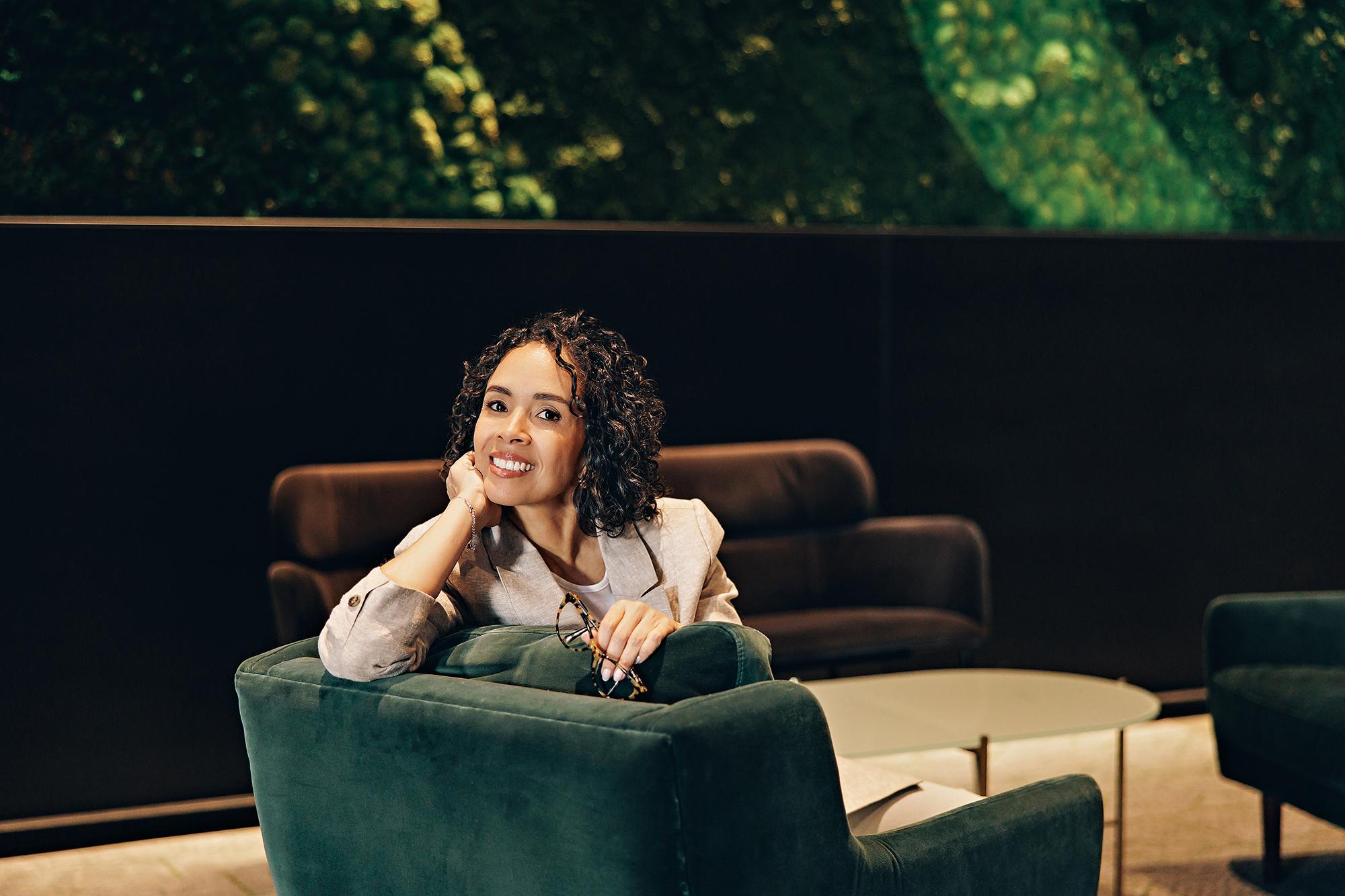 Business headshots Calgary: Female professional posing with hand on chin and holding glasses over a green velvet armchair