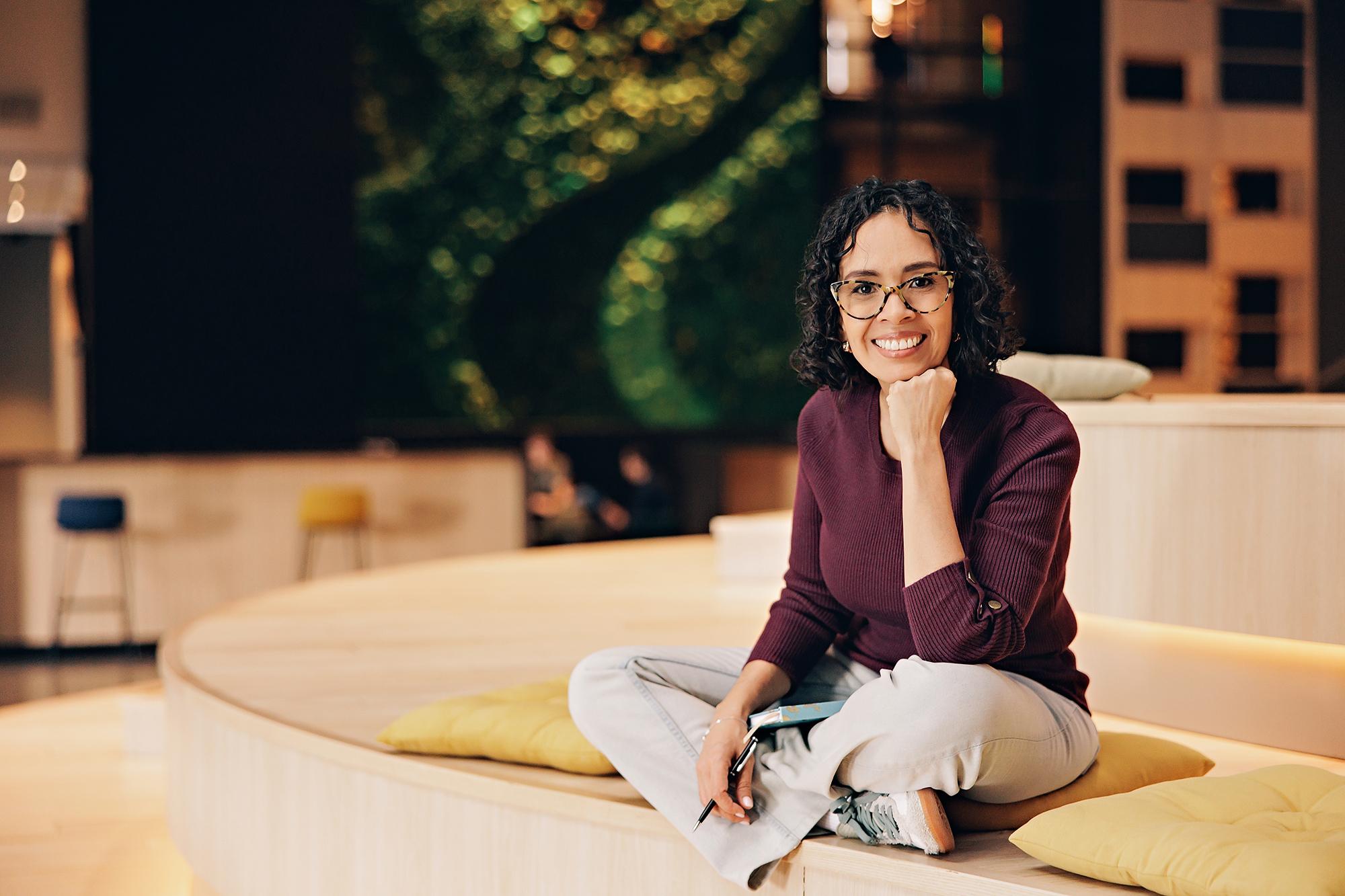 Professional lifestyle headshots in Calgary: Businesswoman in a burgundy sweater resting her chin on her hand and smiling at the camera in a modern workspace