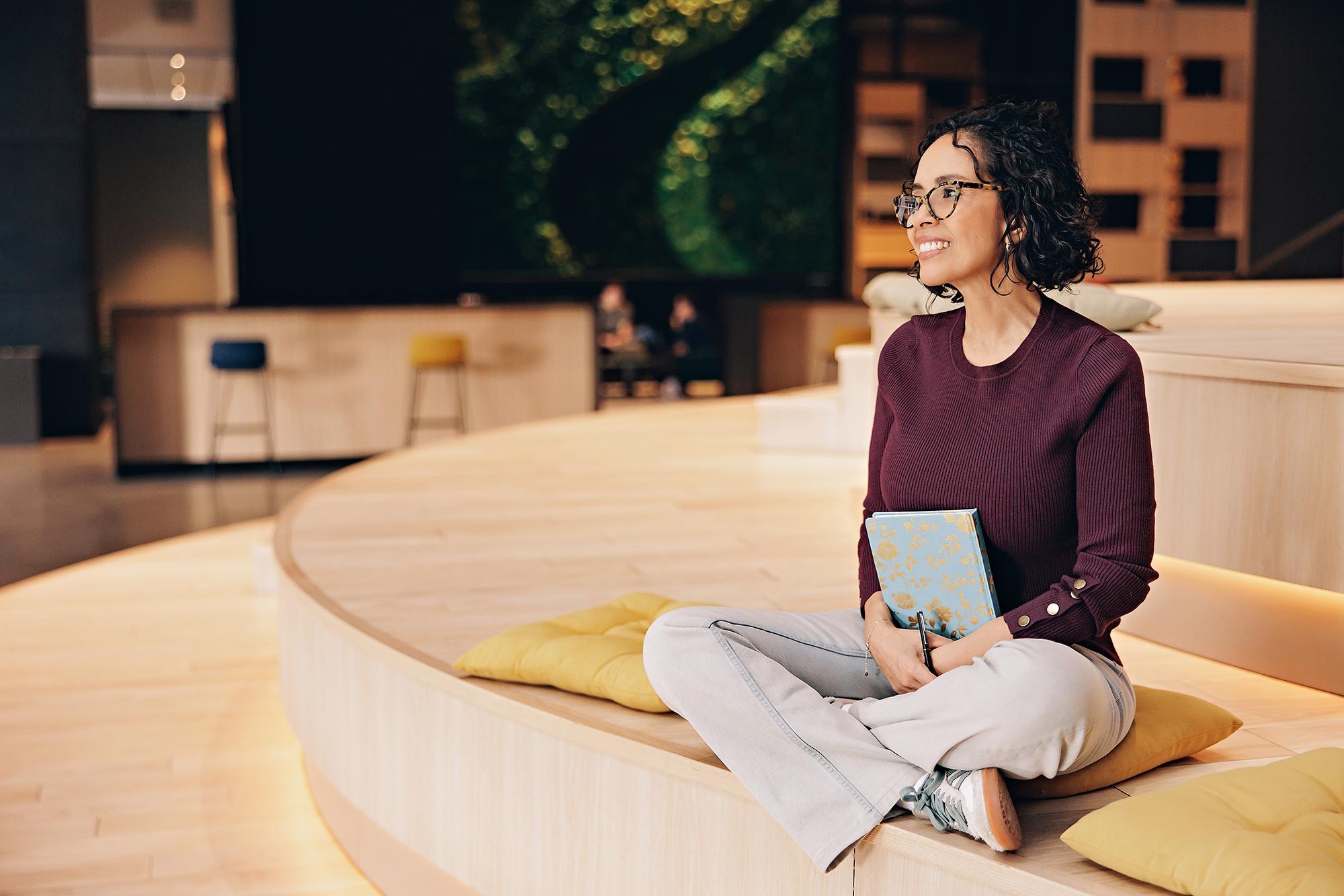 Calgary personal branding photographer: Smiling female entrepreneur in a burgundy sweater sitting cross-legged on wooden tiered seating, holding a floral notebook