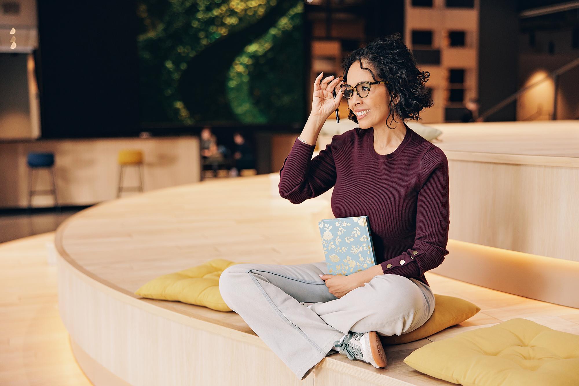 Modern professional headshots Calgary: Smiling businesswoman adjusting her glasses while sitting on wooden tiered seating with a notebook