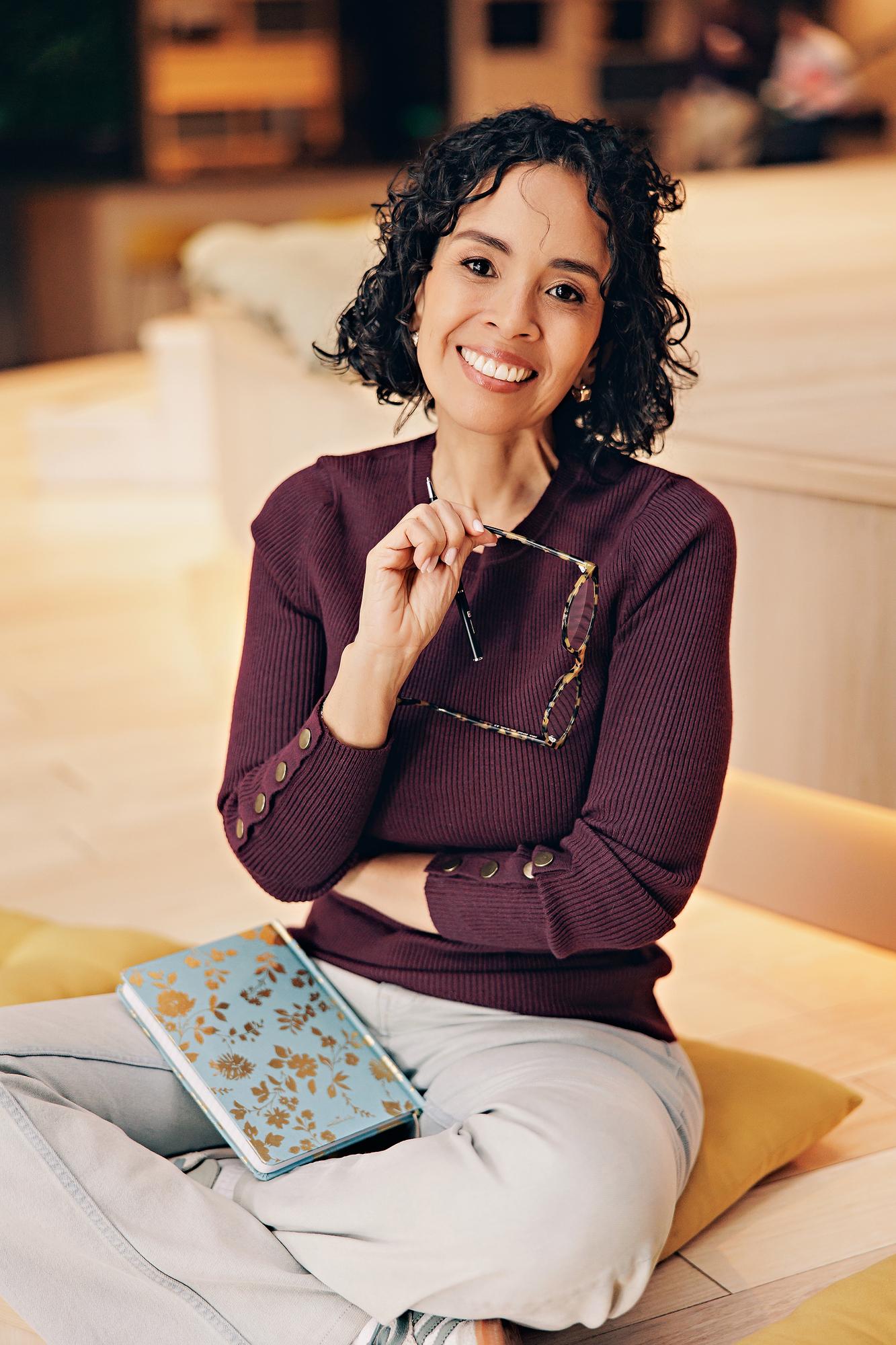 Calgary business portraits: Woman in a burgundy sweater sitting on wooden workspace seating, smiling and holding her tortoiseshell glasses