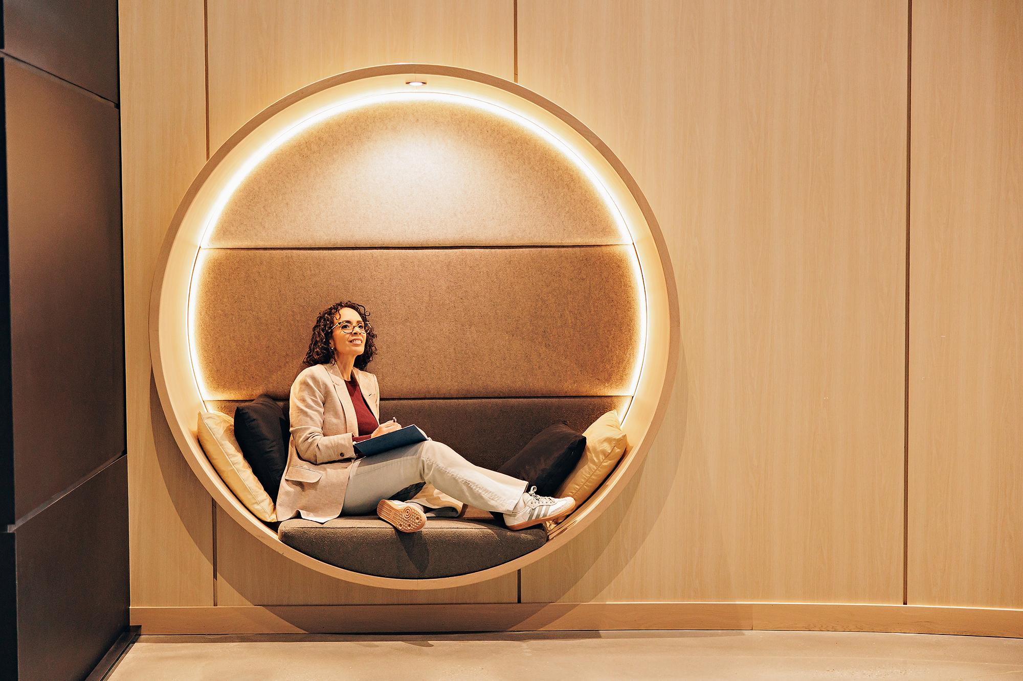 Personal branding photography in Calgary: Smiling businesswoman in a beige blazer sitting inside a modern illuminated circular wall booth, looking thoughtfully away while holding a blue notebook