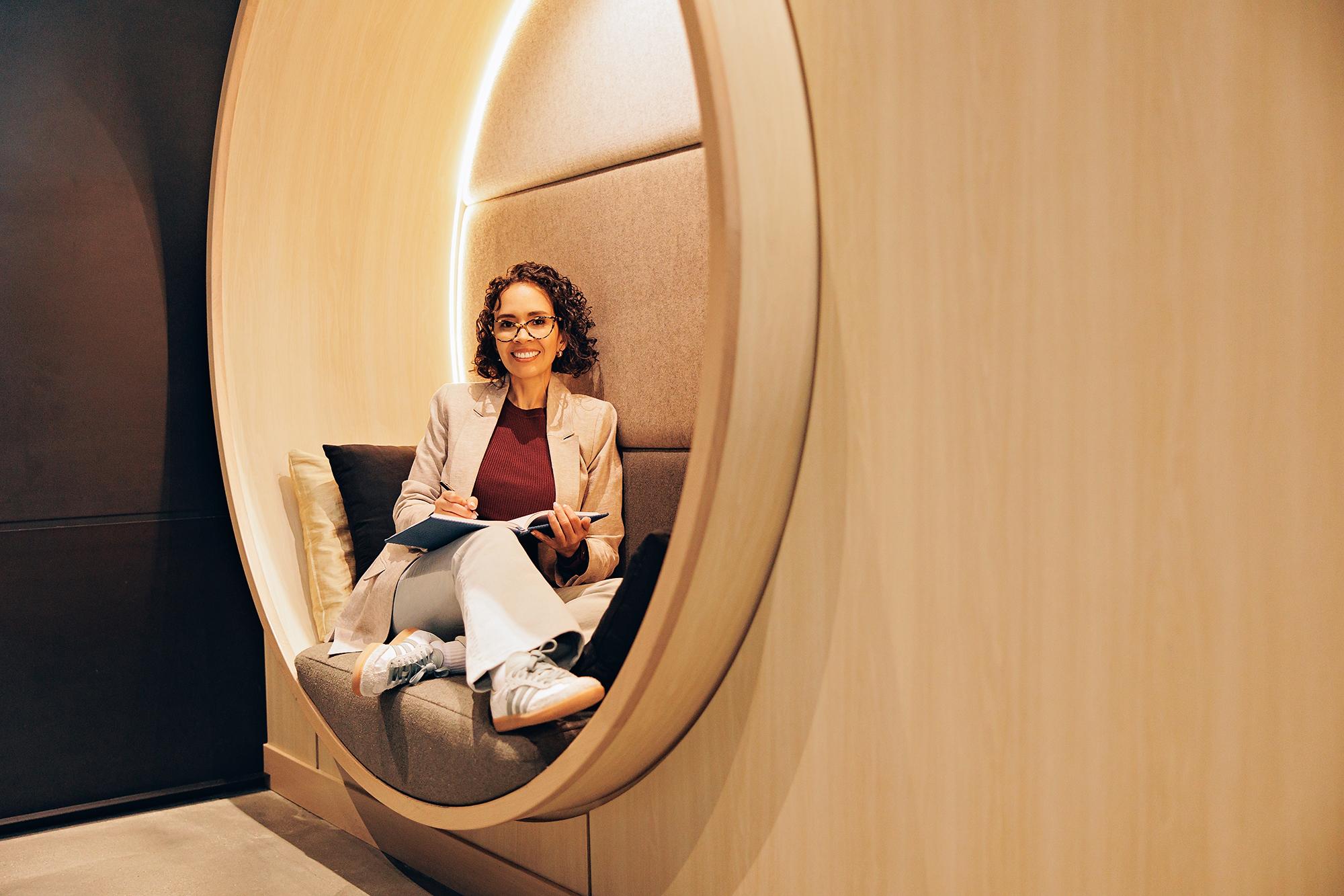 Professional business portraits Calgary: Confident woman in a beige blazer smiling directly at the camera while sitting in a lit circular wall booth with a notebook