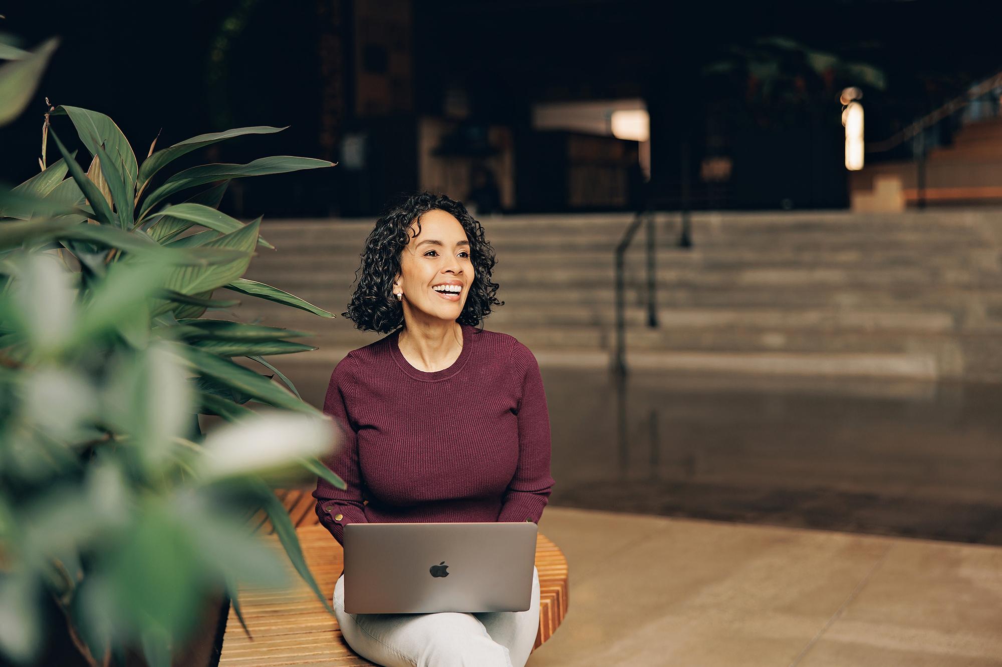 Calgary personal brand photographer: Smiling female entrepreneur in a burgundy sweater sitting on a wooden bench with a laptop, looking away with green plants in the foreground