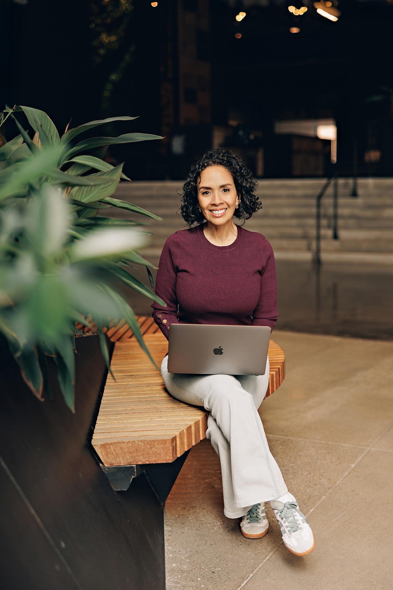 Modern professional headshots Calgary: Businesswoman in a burgundy sweater smiling at the camera while working on a silver laptop on a wooden bench
