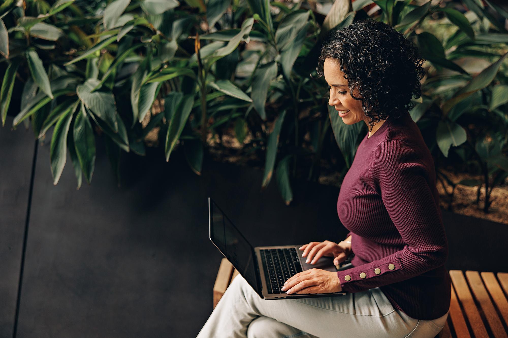 Corporate lifestyle photography Calgary: Female professional typing on a laptop while sitting on a slatted wooden bench next to indoor plants