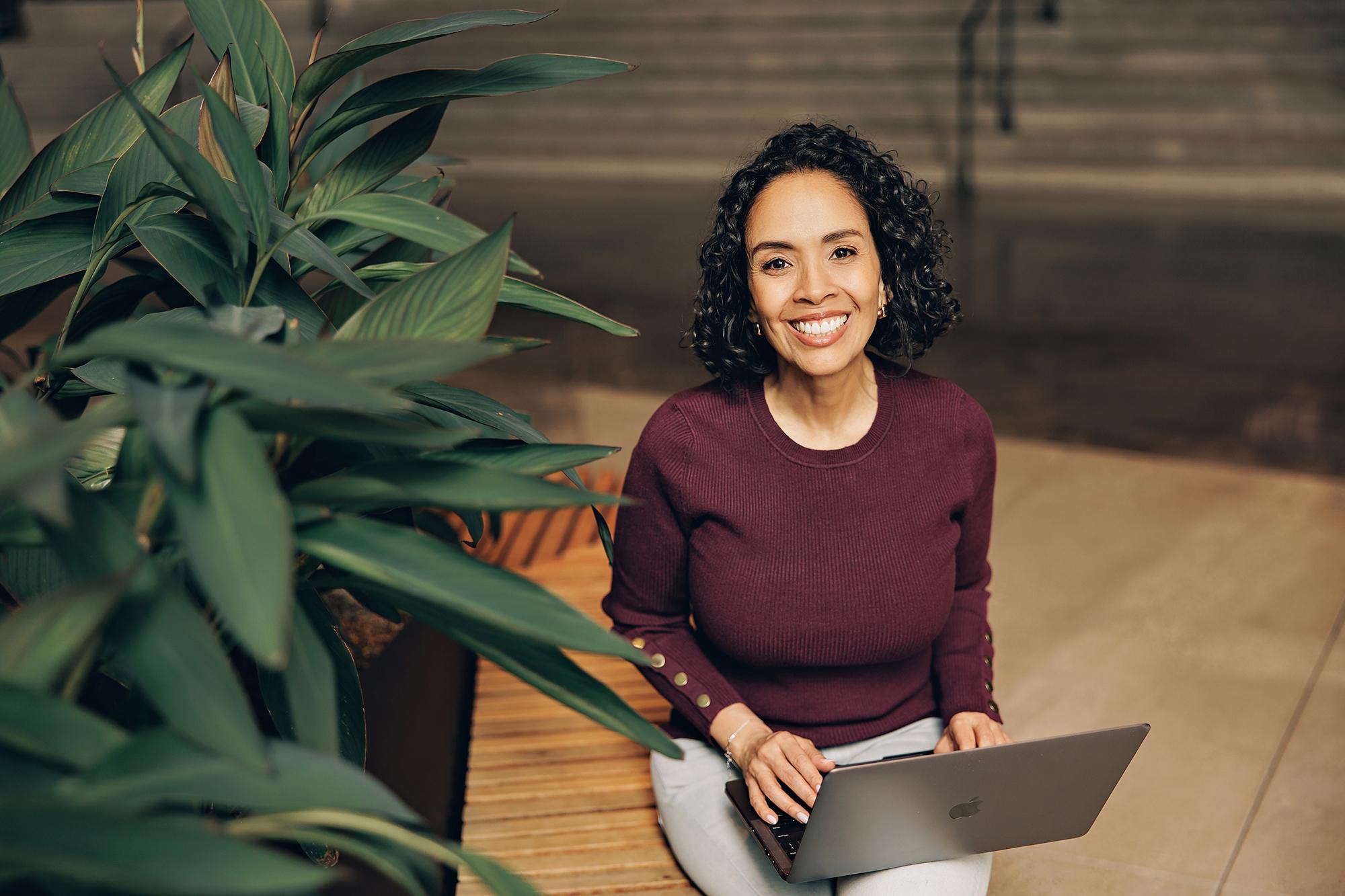 Personal branding session in Calgary: Smiling woman in a burgundy sweater resting her hands on a laptop keyboard while sitting on a wooden indoor bench