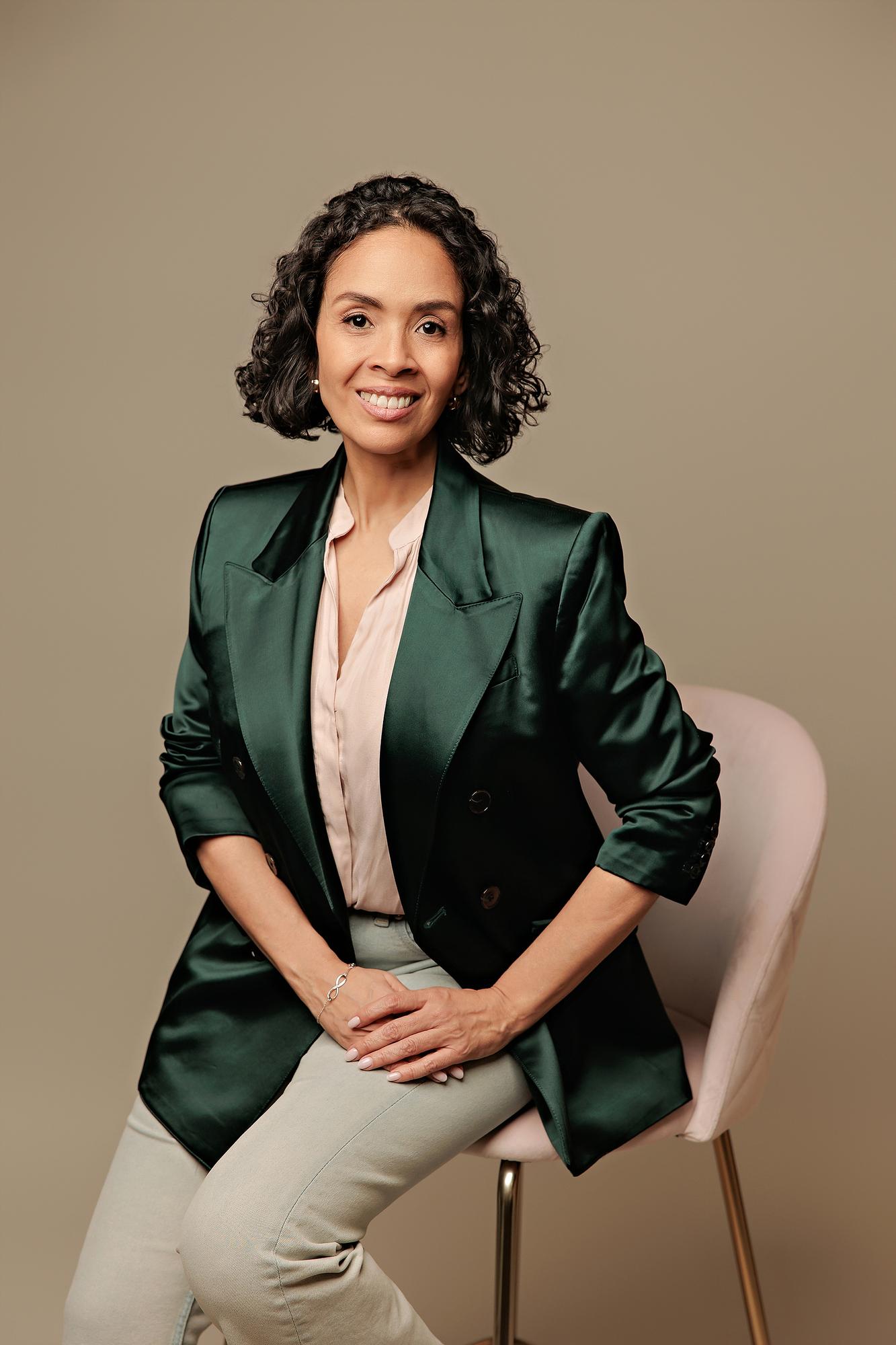 Professional studio headshots Calgary: Confident woman wearing a dark green satin blazer and pink blouse sitting on a pink velvet chair against a neutral beige backdrop